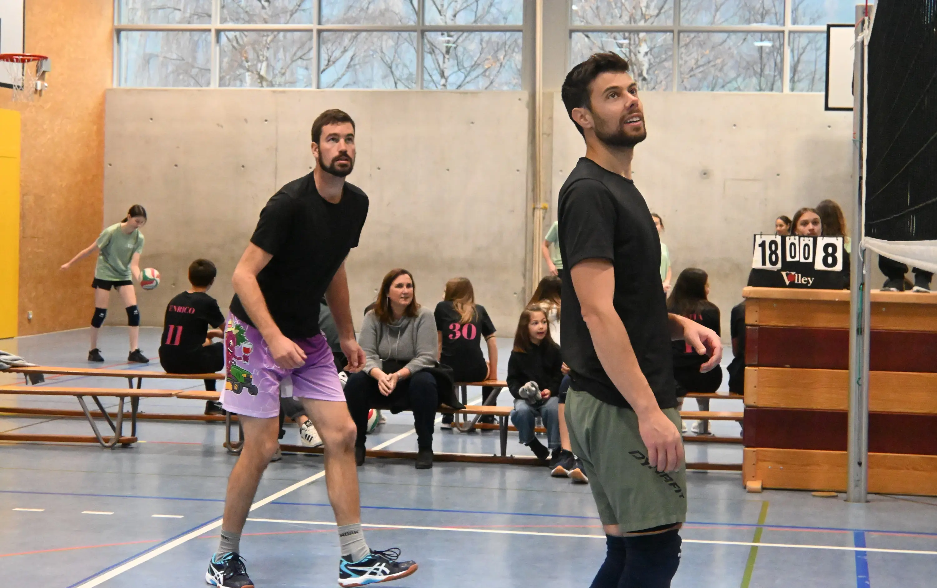 Due giocatori di pallavolo in campo, concentrati nel seguire la palla, mentre nel background si vedono spettatori seduti su panchine.