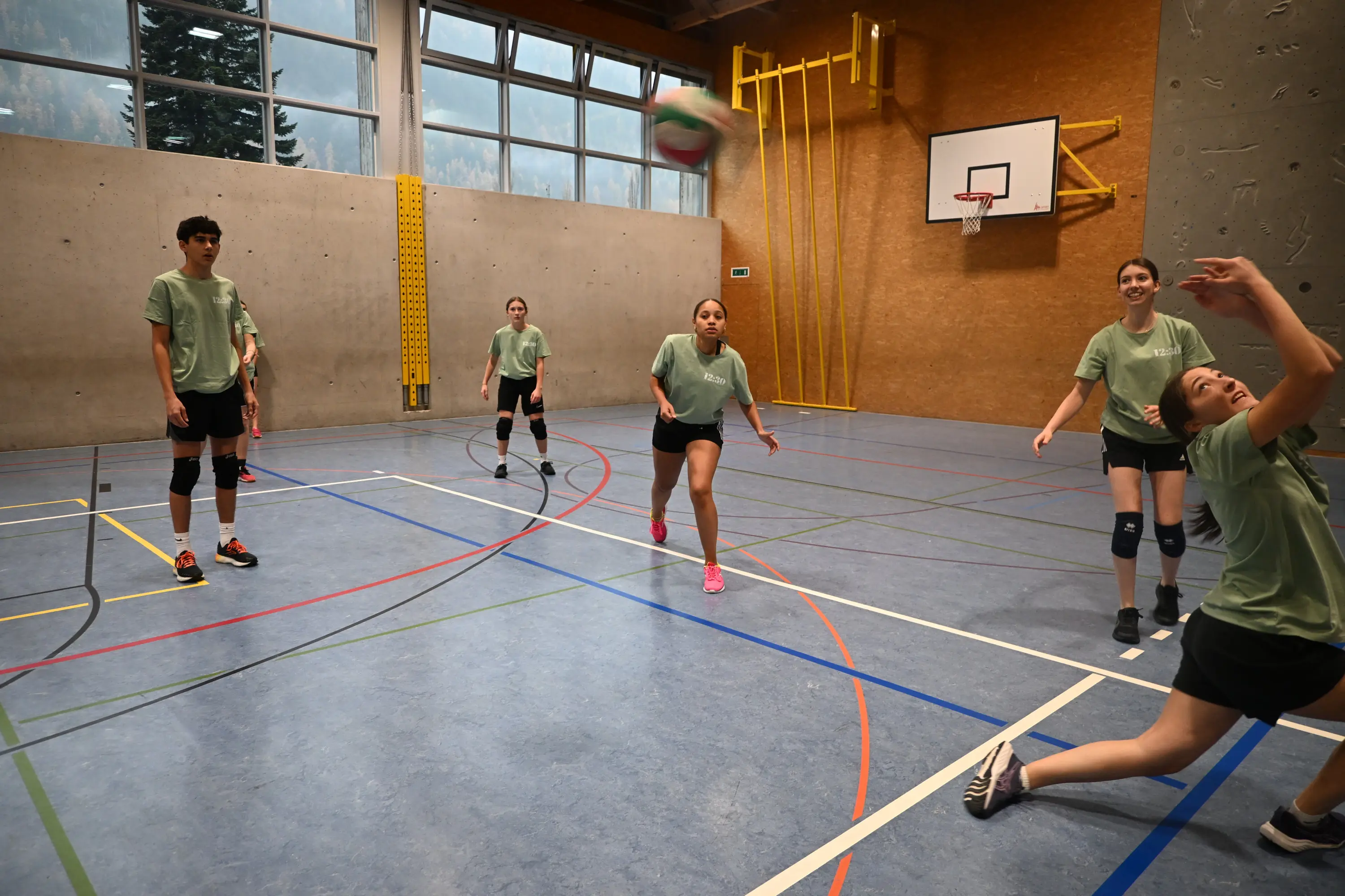 Giovani atleti di pallavolo si allenano in una palestra. Due ragazze colpiscono la palla mentre un ragazzo osserva. Altri due ragazzi sono pronti a ricevere il passaggio.