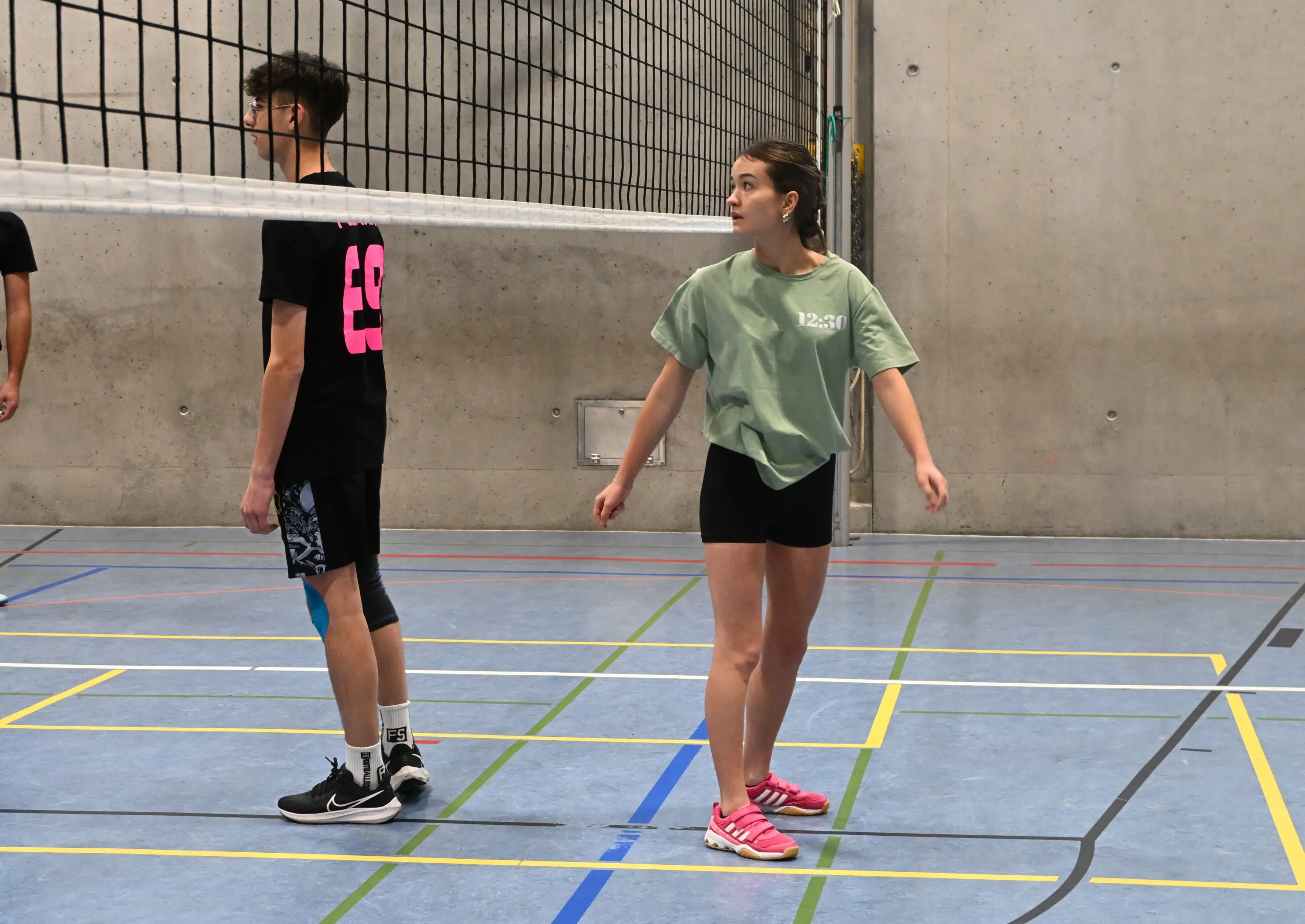 Due giovani giocatori di pallavolo si trovano in un campo da gioco interno. Un ragazzo è girato di spalle, indossando una maglietta nera con un numero rosso, mentre una ragazza indossa una maglietta verde chiaro e pantaloni cort