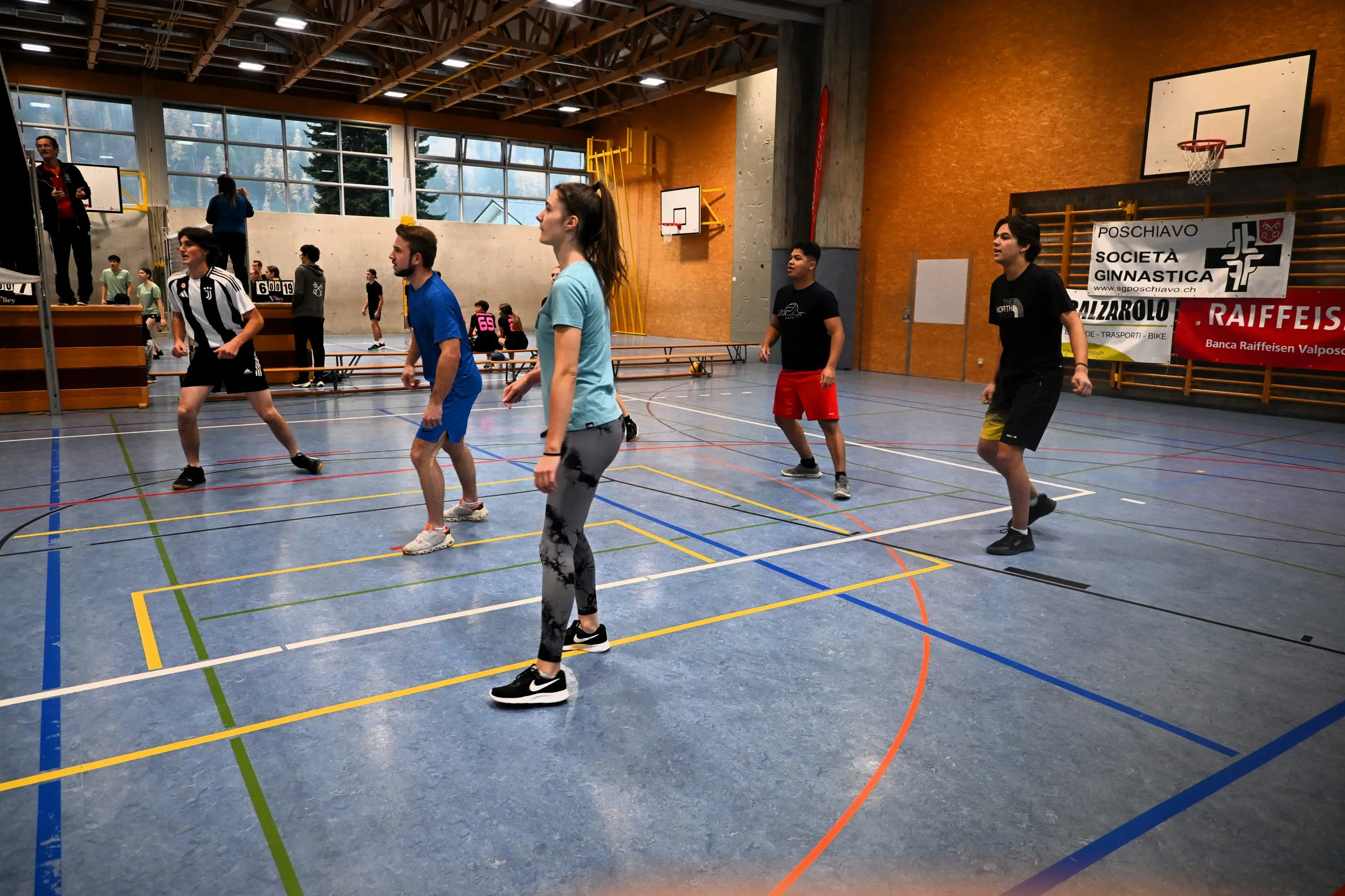 Un gruppo di giovani atleti si prepara a giocare a pallavolo all'interno di un campo sportivo. Sono in diverse posizioni, indossando abiti sportivi colorati. Sullo sfondo si nota una tribuna con alcuni spettatori.