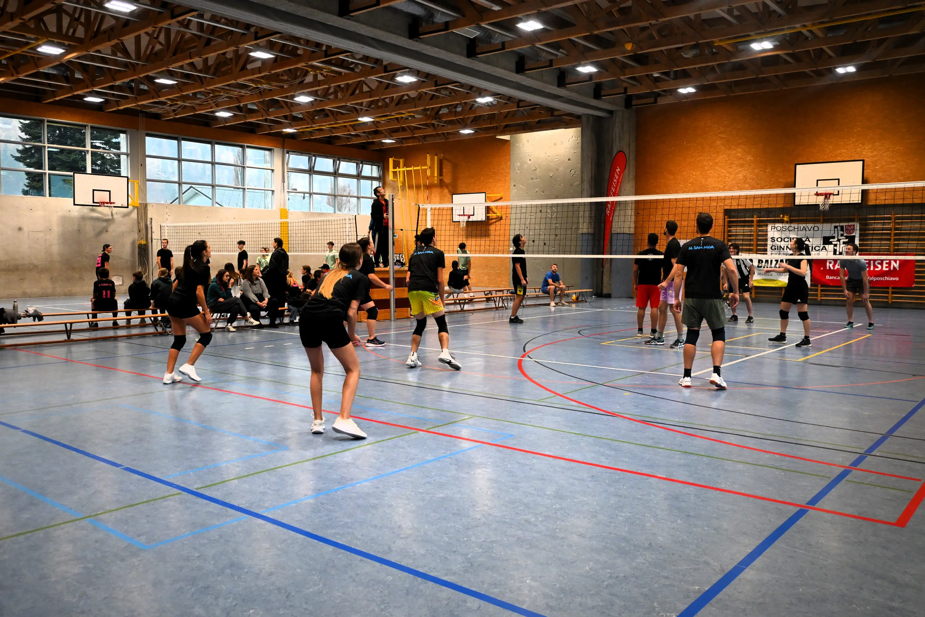Un campo da pallavolo all'interno di una palestra, con giocatori in azione. Alcuni si preparano per un servizio, mentre altri sono in attesa a bordo campo. La struttura è ben illuminata, con canestri di basket visibili sullo sfondo.