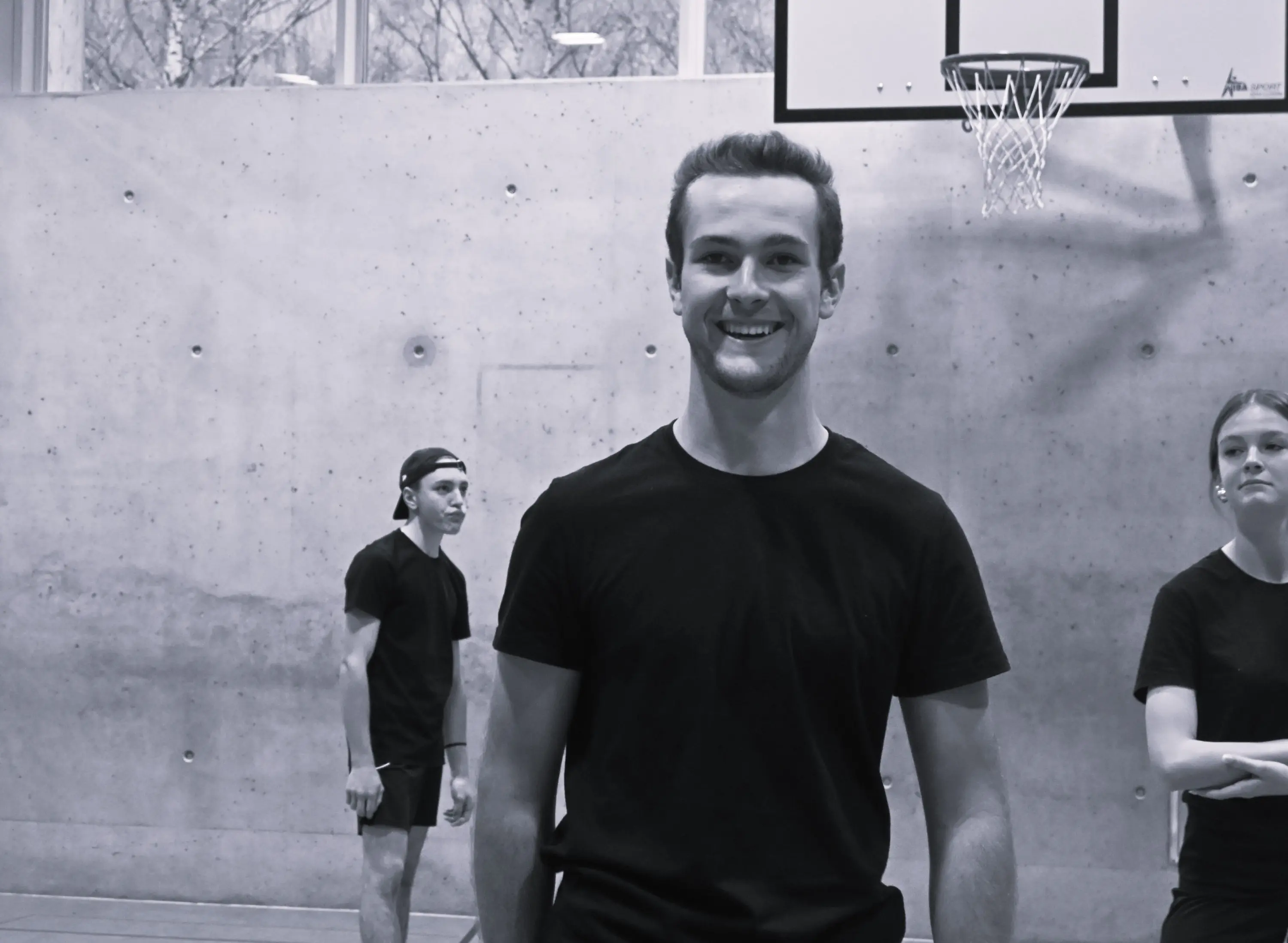 Un giovane sorridente indossa una maglietta nera, mentre sullo sfondo si vedono altre due persone impegnate in un'attività sportiva all'interno di un campo da basket.