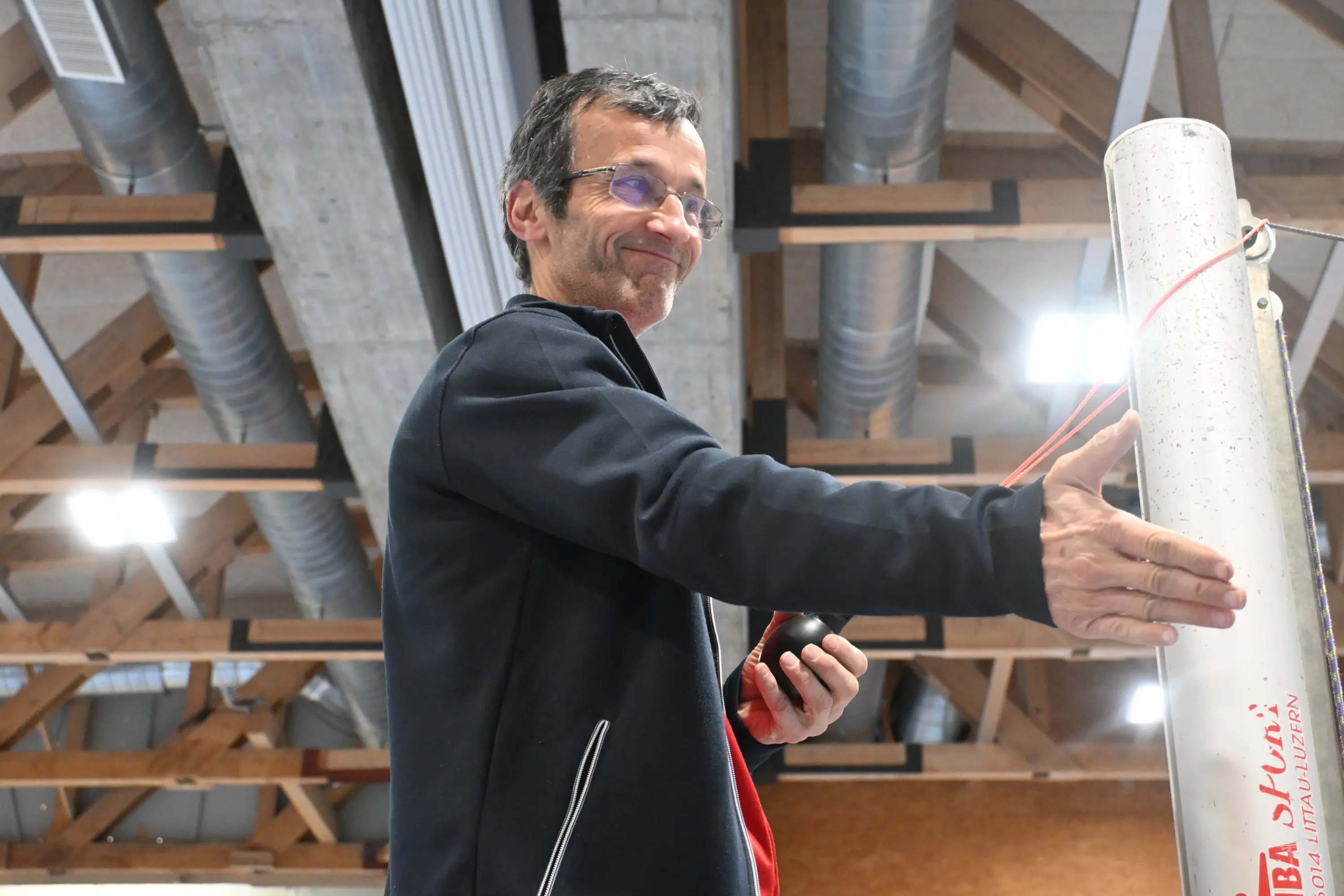 Uomo sorridente che stringe la mano a qualcuno, in un ambiente interno con travi in legno e tubi di metallo visibili.