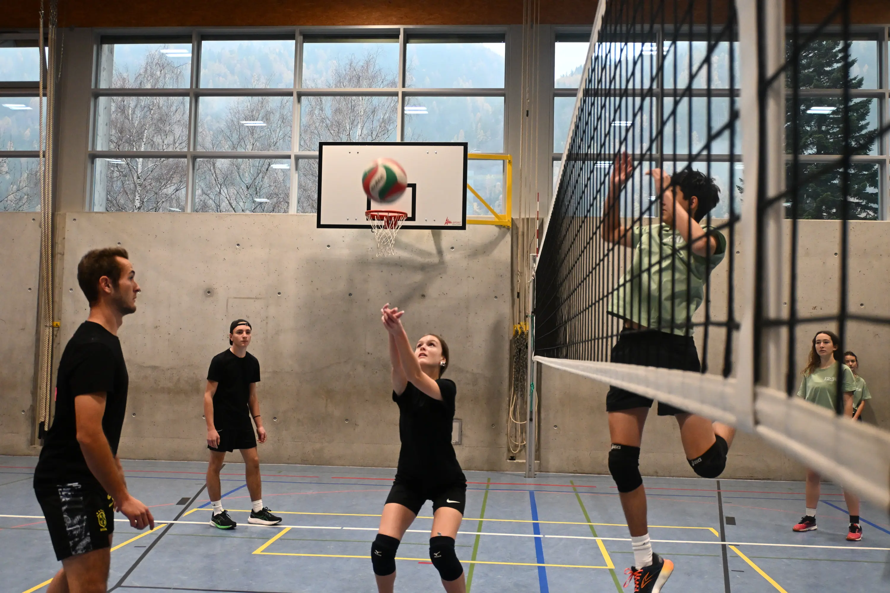 Giovani giocatori di pallavolo in un palazzetto sportivo, uno dei quali sta per colpire la palla mentre gli altri si preparano a riceverla. Una parete di vetro mostra montagne sullo sfondo.