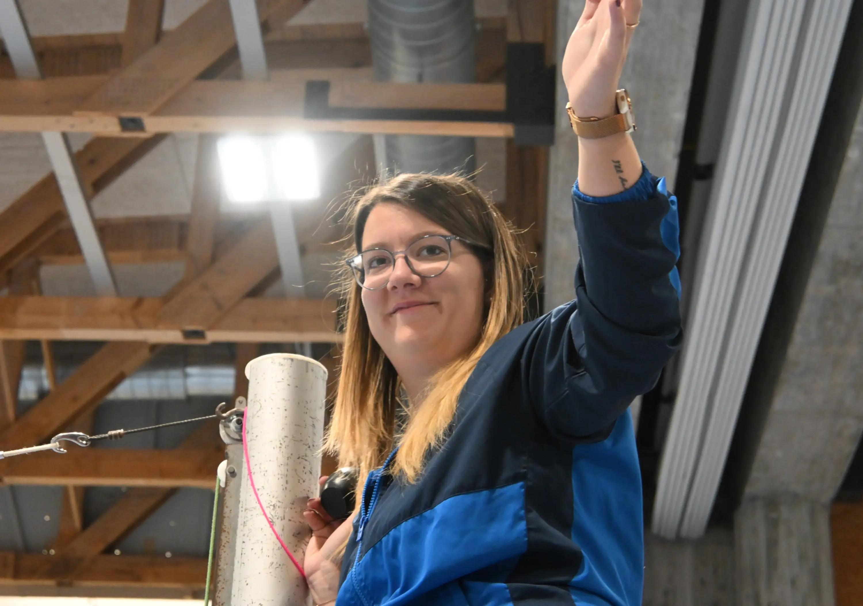 Giovane donna con occhiali e capelli castani, saluta con una mano mentre si trova in un ambiente di lavoro, probabilmente all'interno di un edificio moderno. Indossa una giacca blu e un orologio al polso.