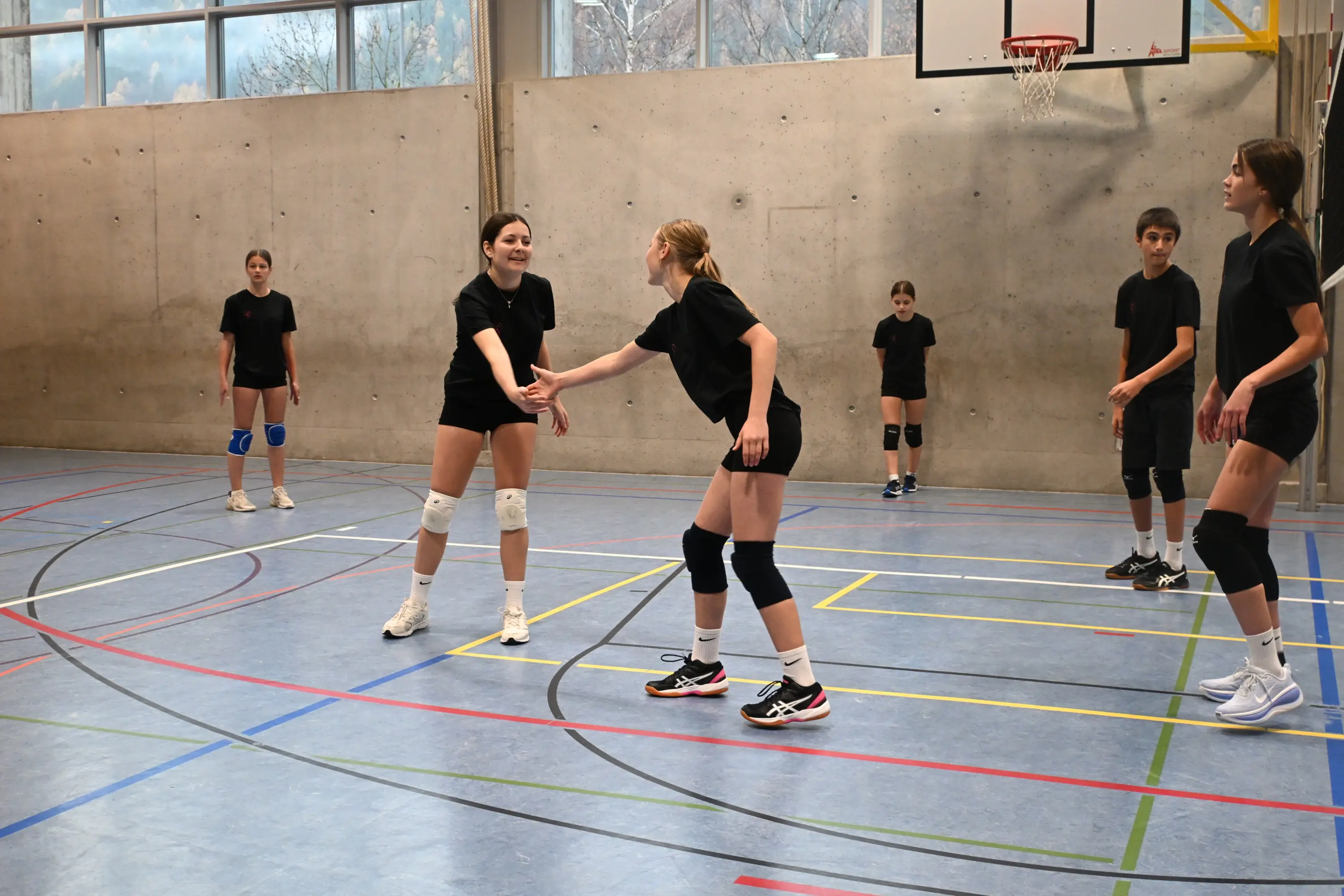 Tre ragazze in maglietta nera e pantaloni sportivi si stringono la mano nel campo di pallavolo. Altre ragazze e un ragazzo sono in attesa sullo sfondo. Il pavimento è blu con linee colorate.