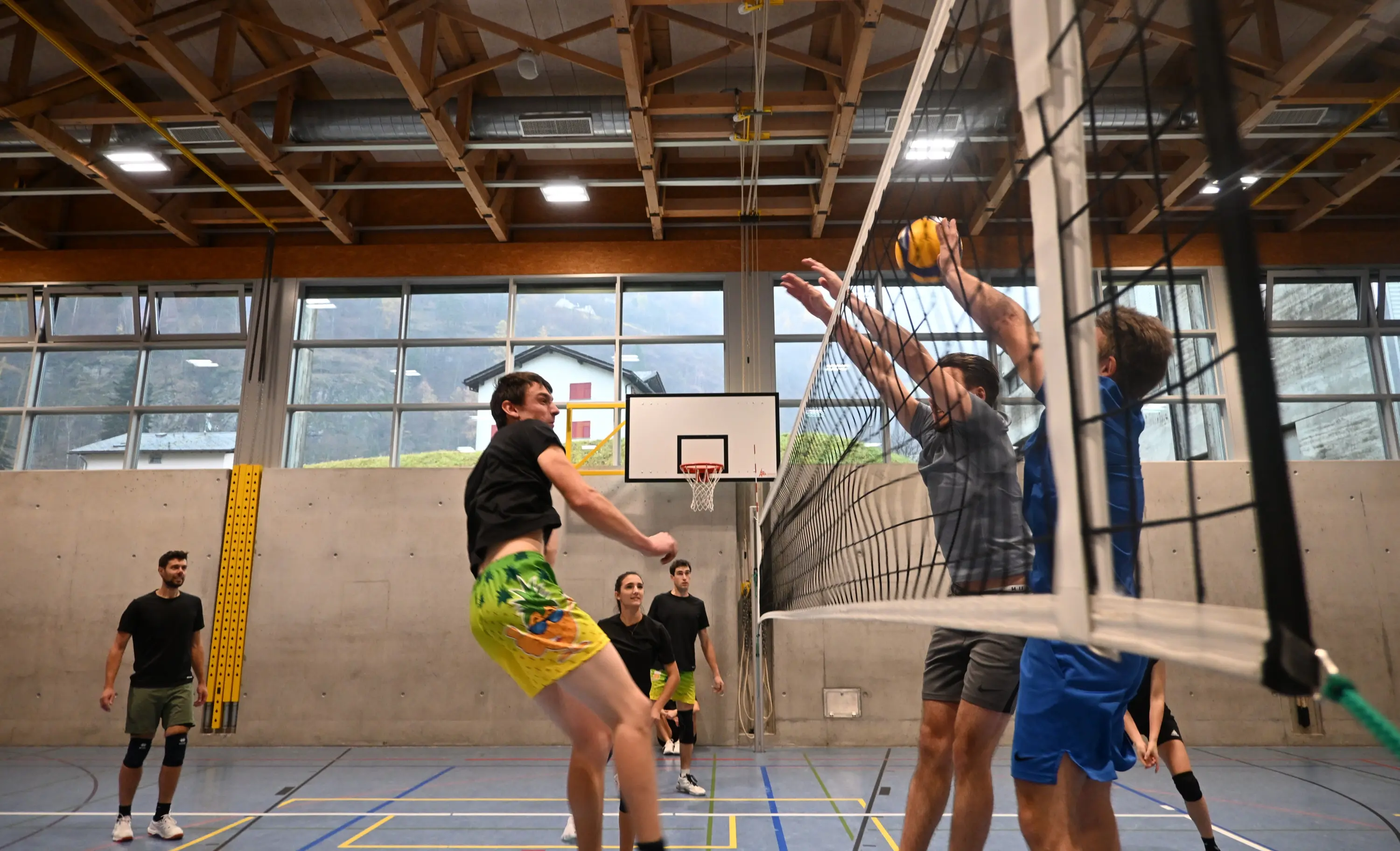 Giocatori di pallavolo in azione in una palestra. Un atleta colpisce la palla mentre altri due tentano di effettuare il blocco. Sullo sfondo si vedono altri giocatori e una grande finestra con vista su colline.