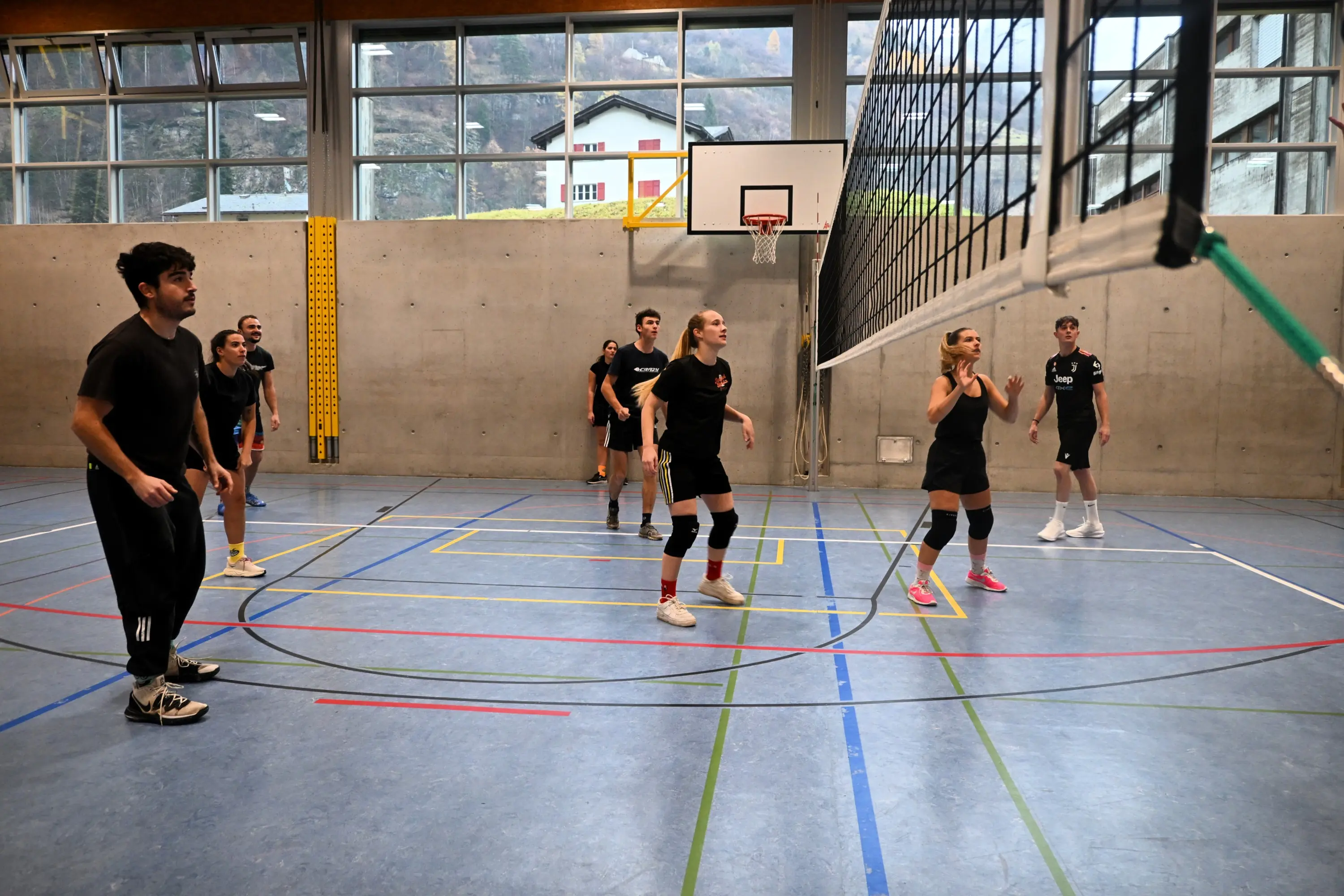 Gruppo di giovani atleti che giocano a pallavolo in un campo all'interno di una palestra con pareti grigie e finestre grandi.