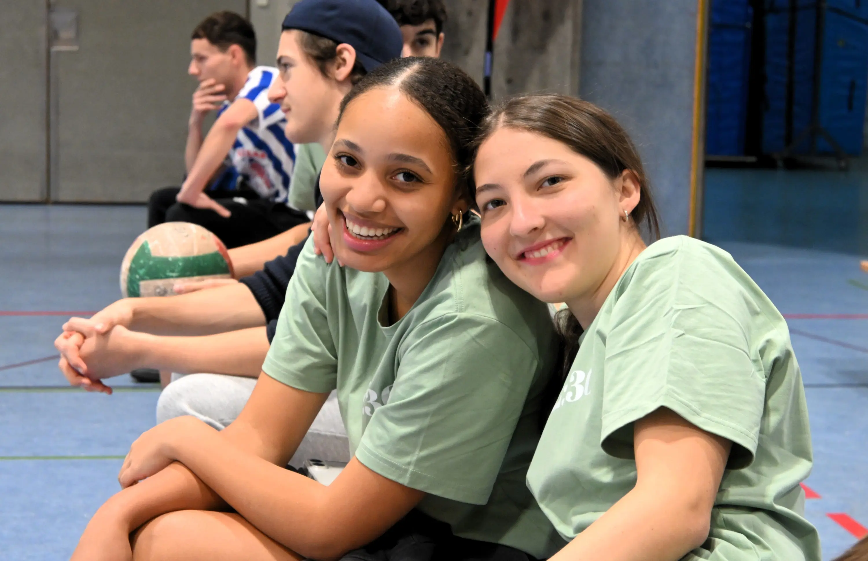 Due ragazze sorridenti in magliette verdi si abbracciano mentre siedono su una panchina, circondate da altri giovani. Sullo sfondo si intravede una palestra con attrezzi sportivi.