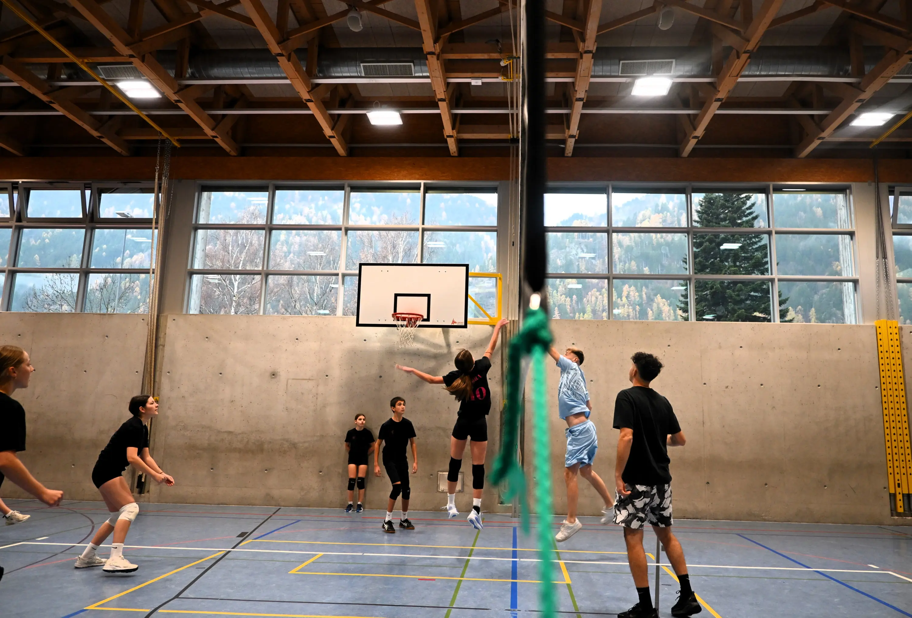 Un gruppo di giovani giocatori di pallavolo in azione in una palestra. Un giocatore salta per effettuare un attacco mentre gli altri si preparano per la ricezione. Grandi finestre mostrano un paesaggio montano sullo sfondo.