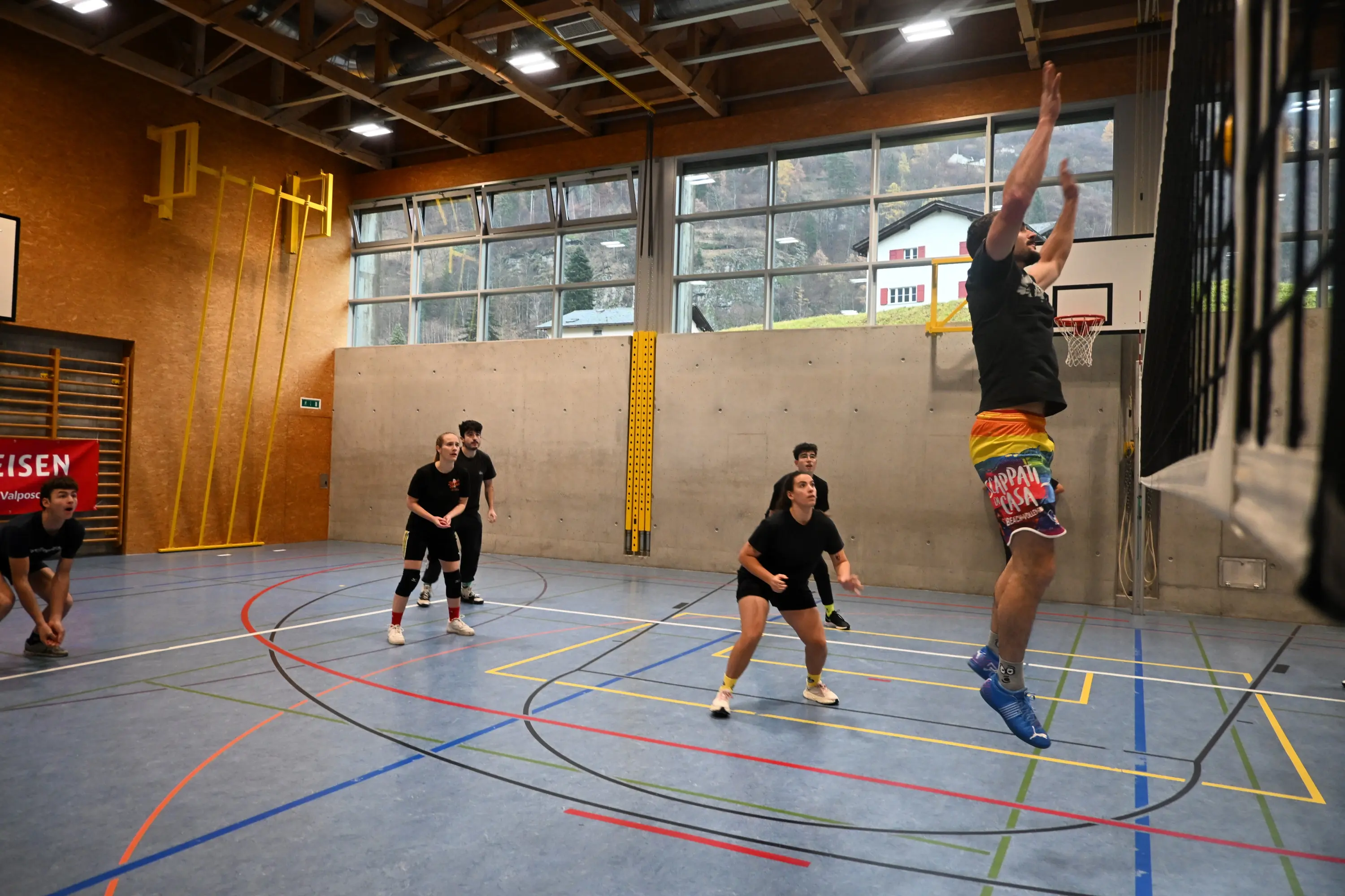 Giocatori di pallacanestro in un campo al coperto, mentre uno di loro salta per fare un canestro, mentre gli altri si preparano a ricevere la palla. Il campo è delimitato da linee colorate e ci sono finestre che illuminano l'ambiente