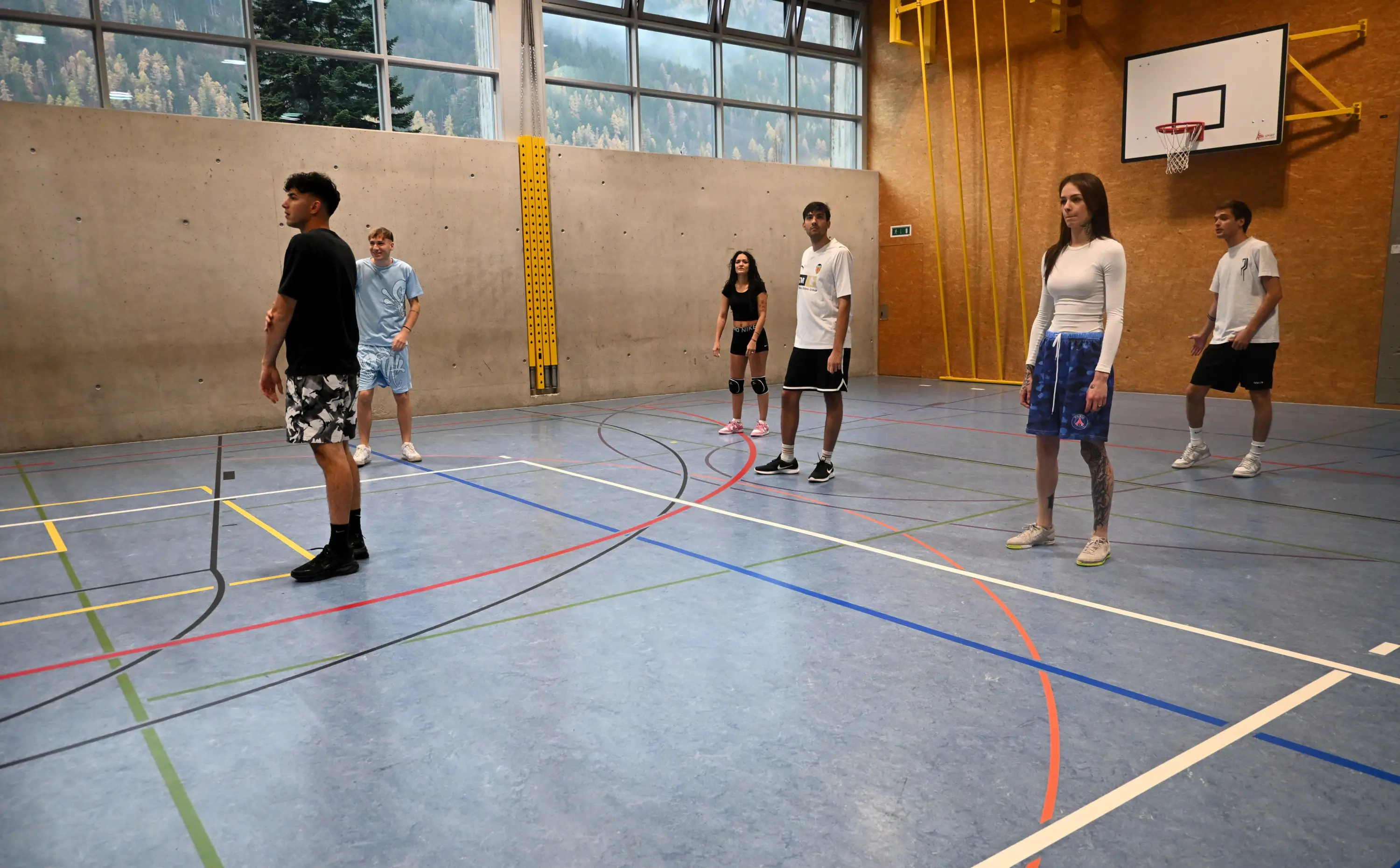 Un gruppo di sei giovani in una palestra, alcuni indossano abbigliamento sportivo mentre si preparano a giocare a pallone. Le pareti sono di cemento e c'è un canestro da basket sullo sfondo.