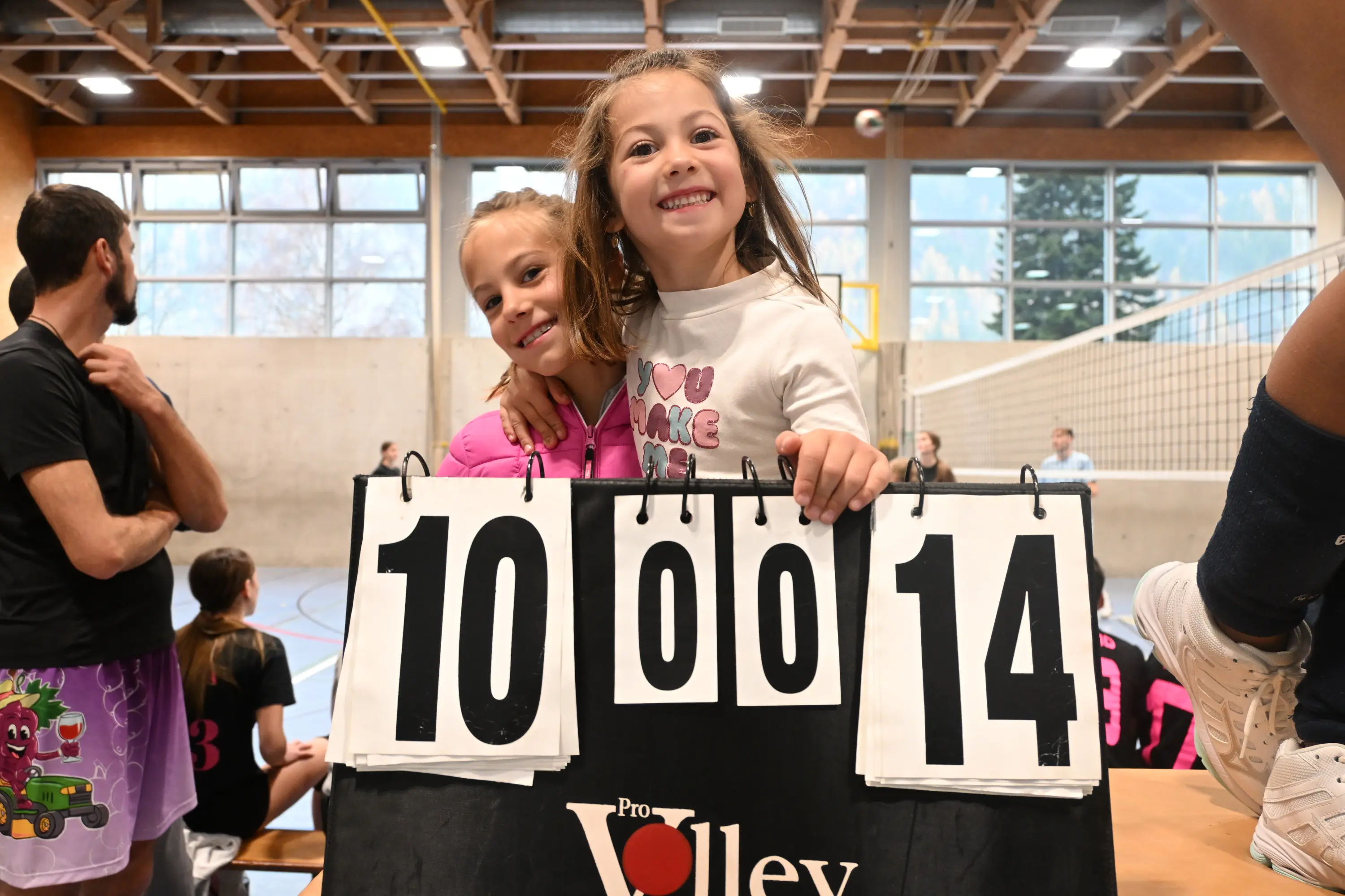 Due ragazze sorridenti abbracciate davanti a un tabellone segnapunti con i numeri 10, 0 e 14. Sullo sfondo si intravedono altri giocatori di pallavolo.