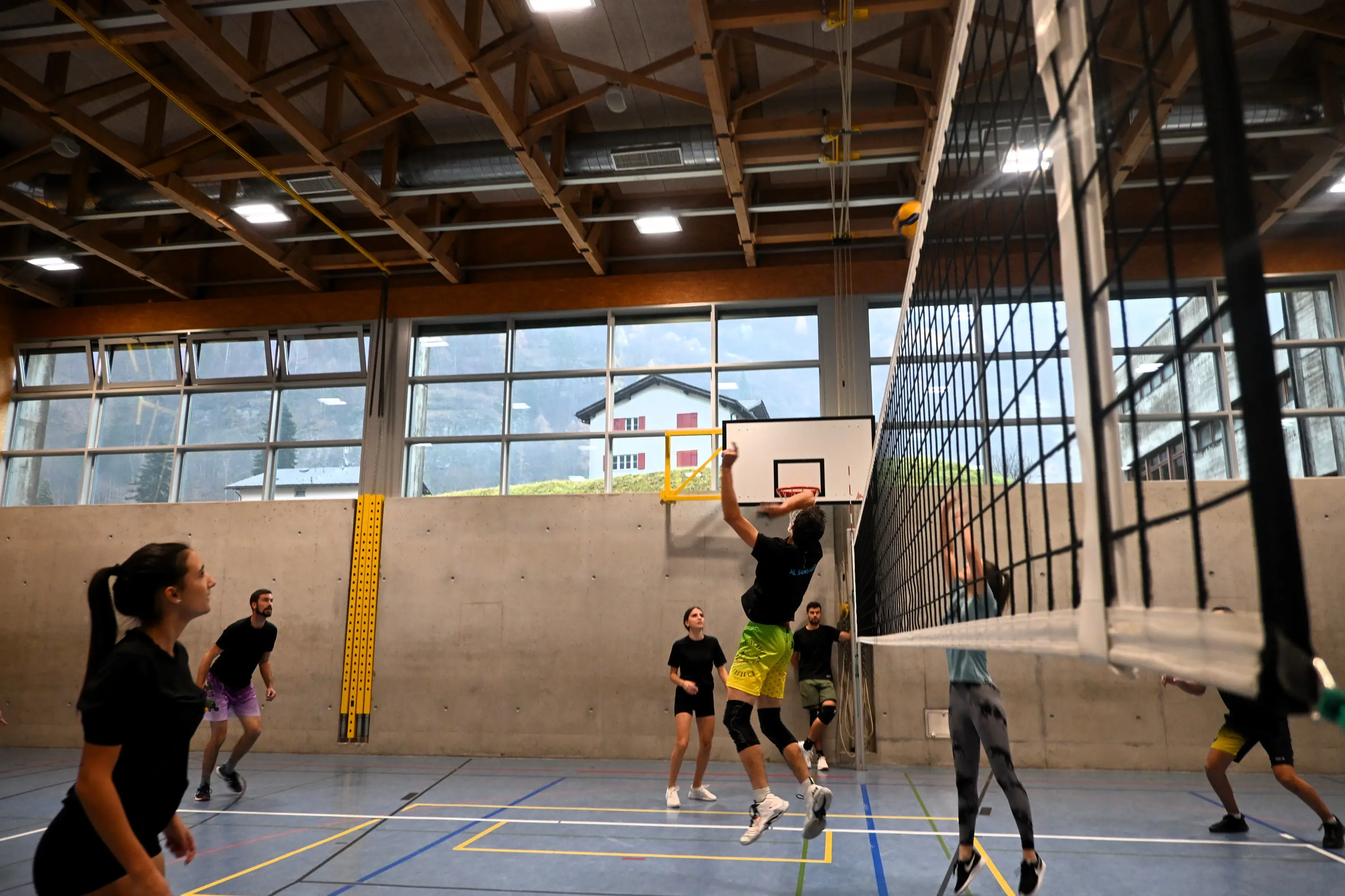 Un gruppo di giocatori di pallavolo in azione in una palestra. Un giocatore sta saltando per colpire la palla mentre gli altri osservano. Sullo sfondo si vedono finestre grandi e una rete da pallavolo.