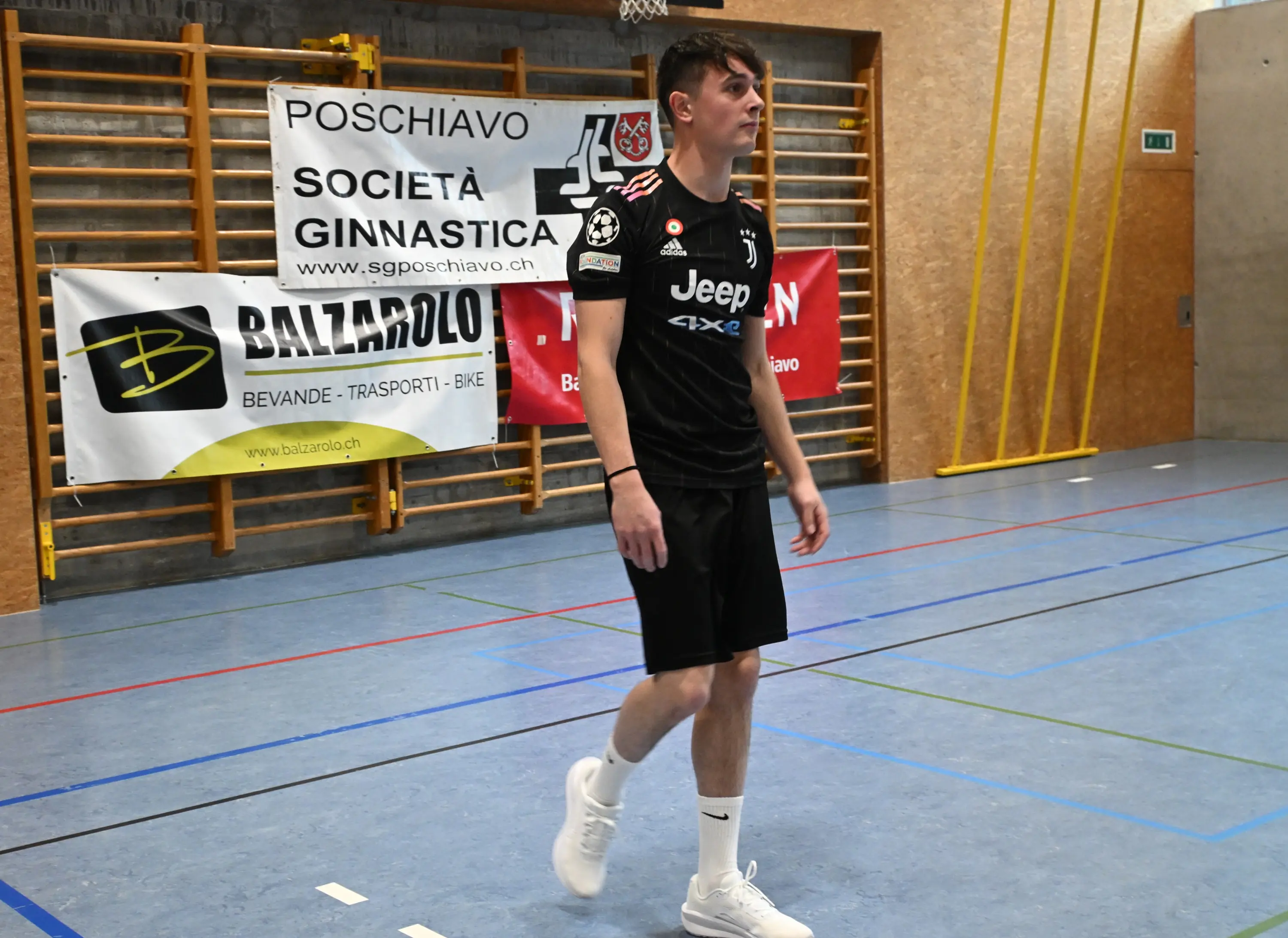 Un giovane atleta in maglia nera con logo Jeep cammina su un campo di pallamano, circondato da bandiere e striscioni di sponsorizzazione.