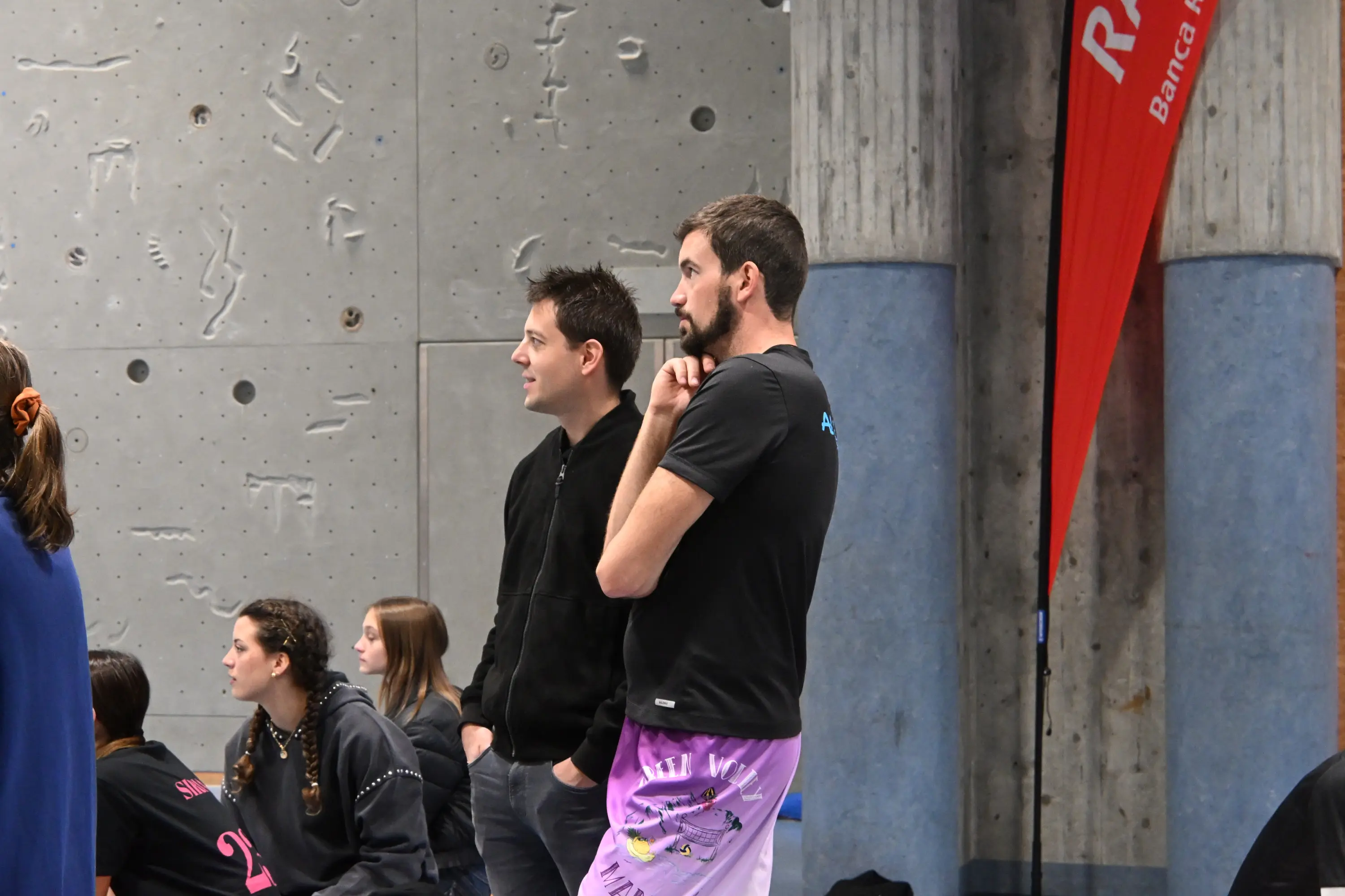 Due uomini osservano un evento in una palestra. Uno indossa una maglietta nera e l'altro una maglietta nera con pantaloni corti rosa. Sullo sfondo, si vedono altre persone e una bandiera rossa.