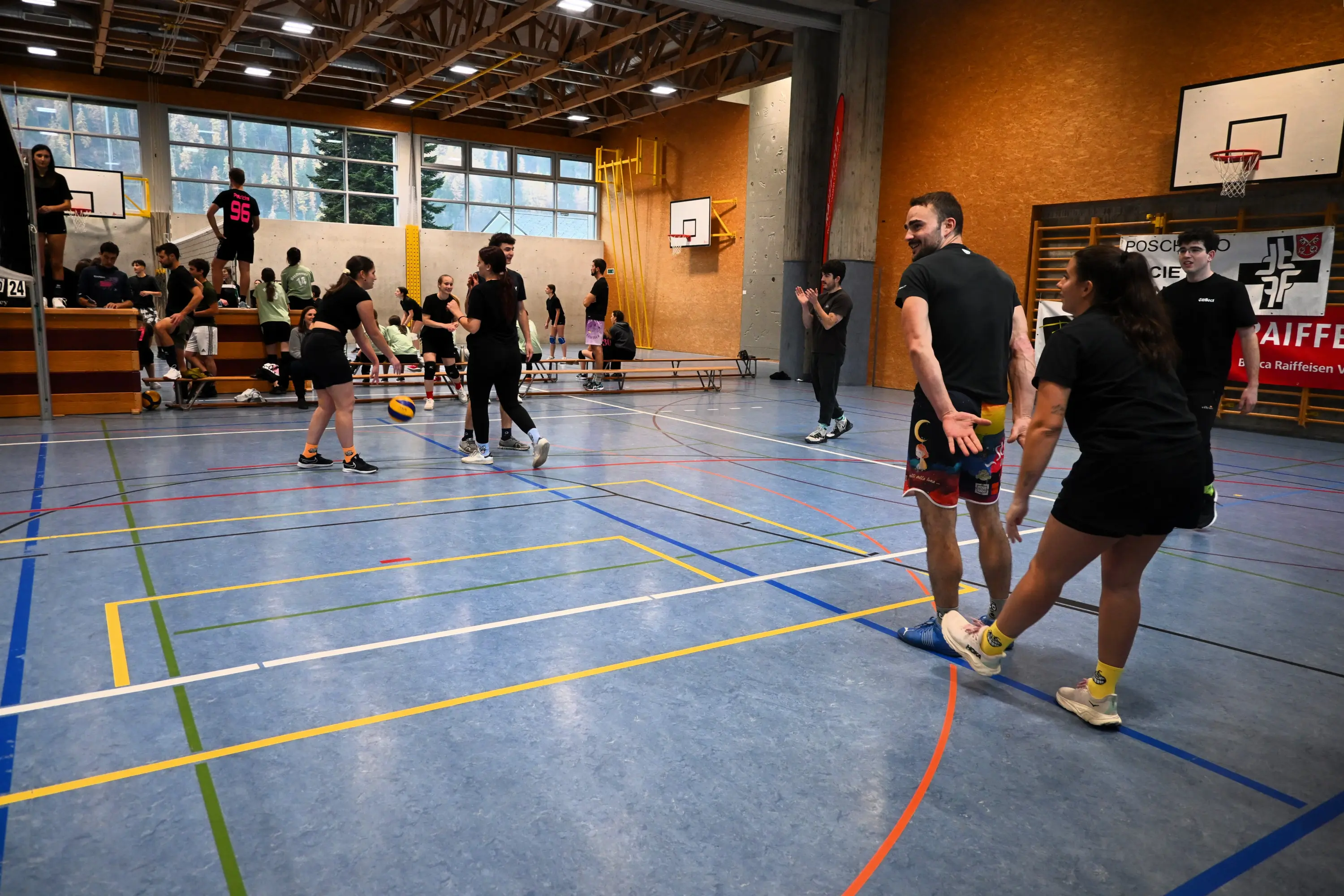 Un gruppo di giovani gioca a pallavolo in un palazzetto sportivo. Sono visibili palloni, reti e attrezzature sportive. Alcuni ragazzi si preparano a ricevere la palla, mentre altri si muovono sul campo.