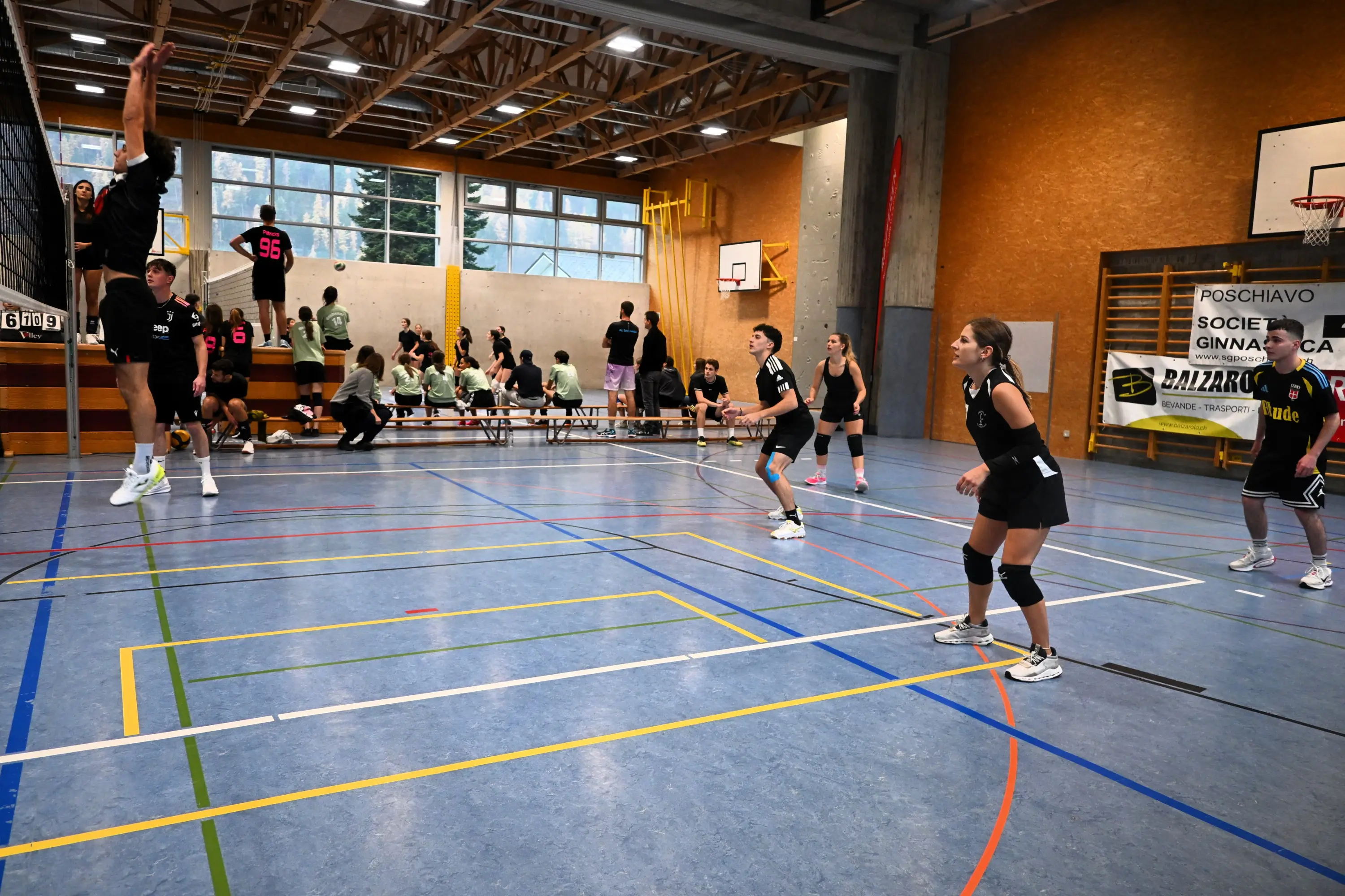 Un gruppo di ragazzi e ragazze in un campo di pallavolo al chiuso. Un giovane sta saltando per colpire la palla, mentre altri giocatori stanno pronti in posizione. In fondo, un pubblico osserva e una lavagna segna il punteggio.