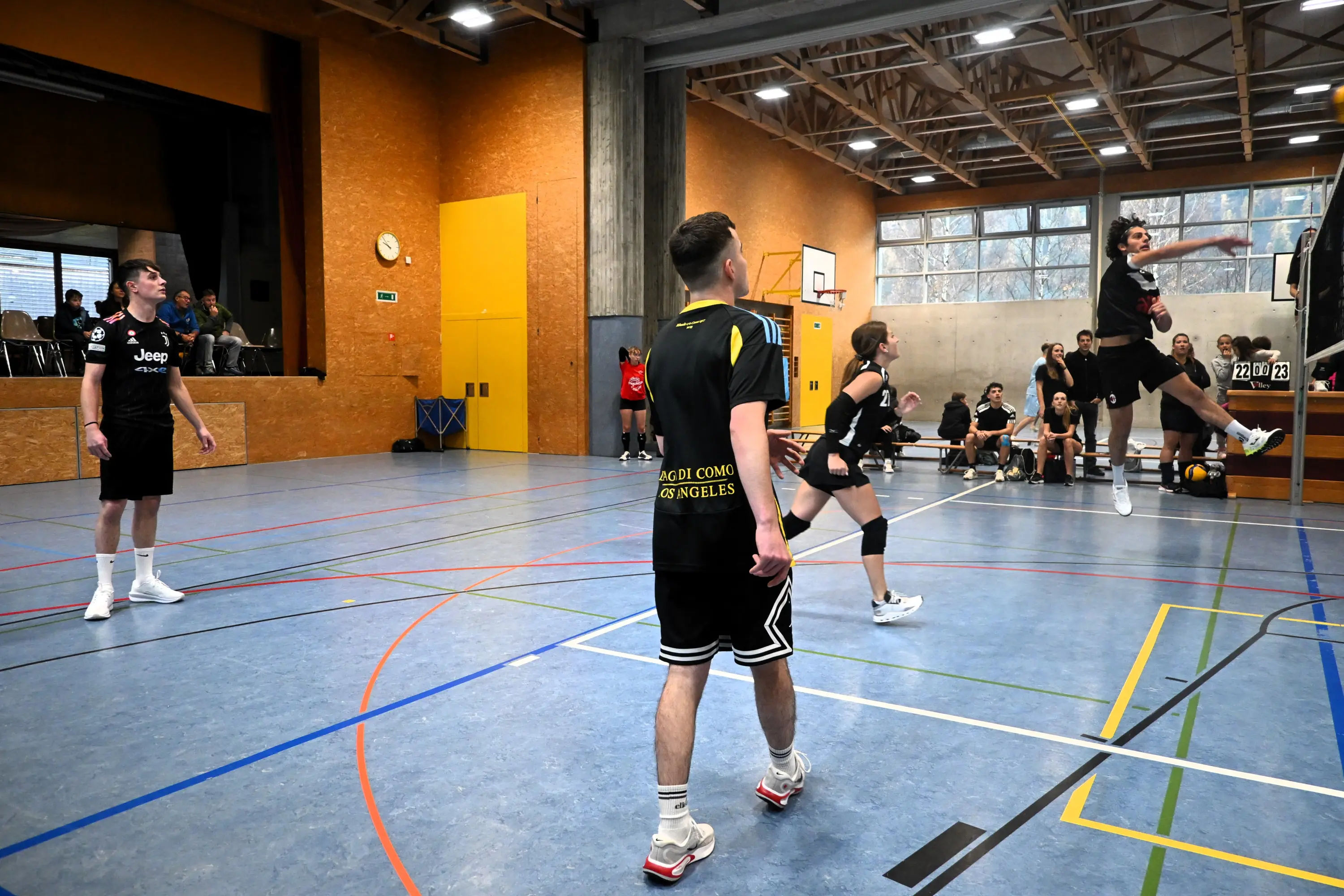 Giocatori di pallavolo in un palazzetto sportivo, mentre uno di loro salta per colpire la palla. Altri giocatori osservano e il pubblico è visibile sullo sfondo.