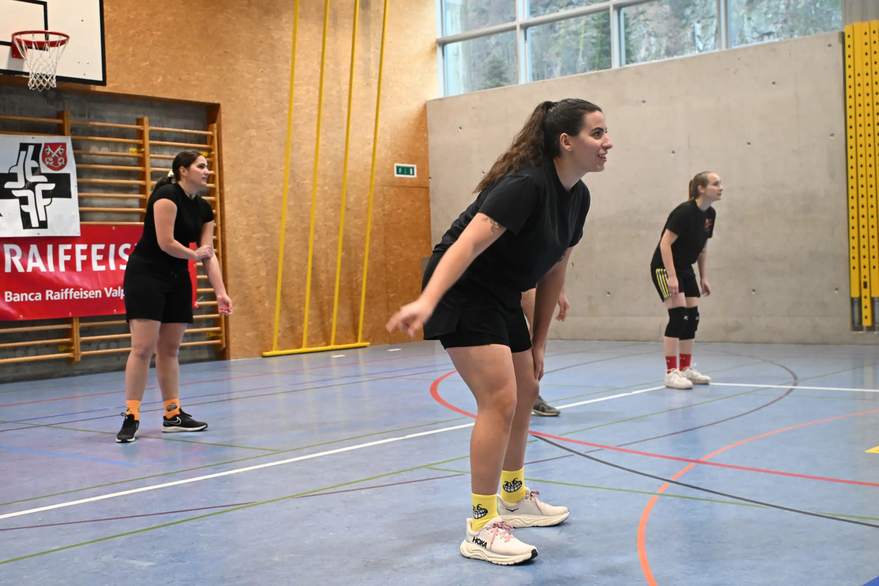 Tre giocatrici di pallacanestro in una palestra, pronte per un'azione. Due di loro indossano magliette nere e pantaloni corti, mentre una ha una postura di attesa. Sullo sfondo, il canestro e una bandiera di sponsorizz