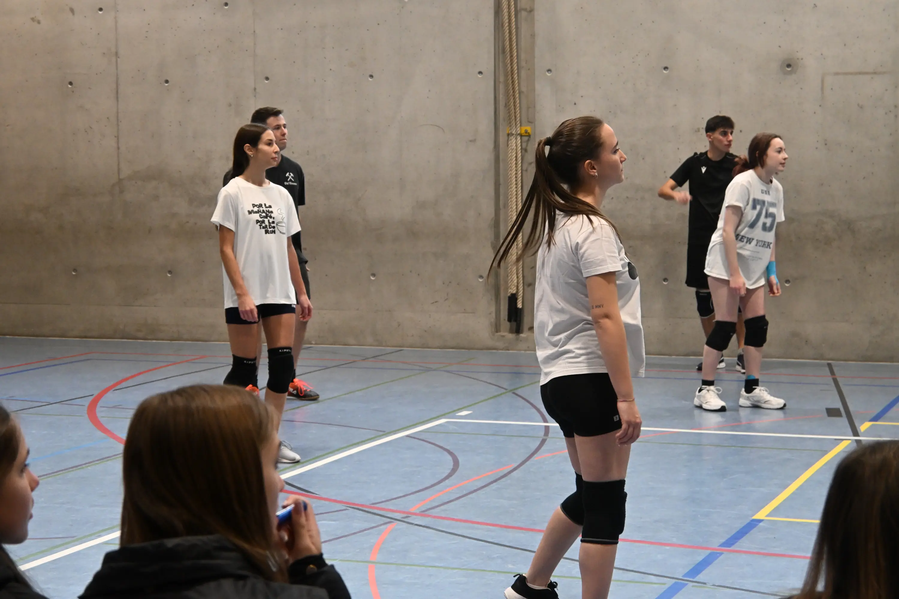 Giovani atleti di pallavolo in una palestra, alcuni in attesa mentre altri si preparano per il gioco. Indossano magliette bianche e pantaloni da sport. Un ambiente sportivo con linee colorate sul pavimento.