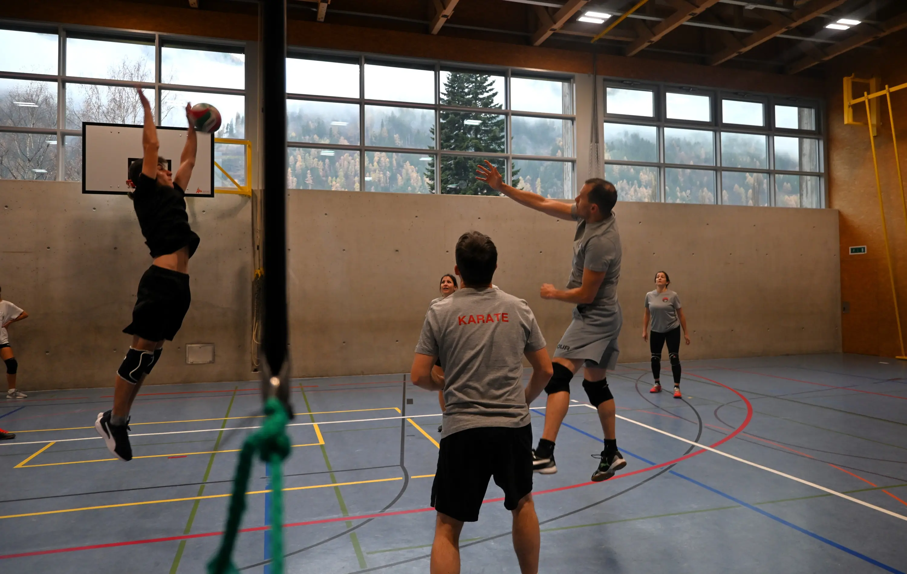 Giocatori di pallavolo in un campo indoor mentre un atleta salta per colpire la palla. Altri giocatori si preparano a ricevere il pallone. Sullo sfondo si vedono finestre con una vista montuosa.