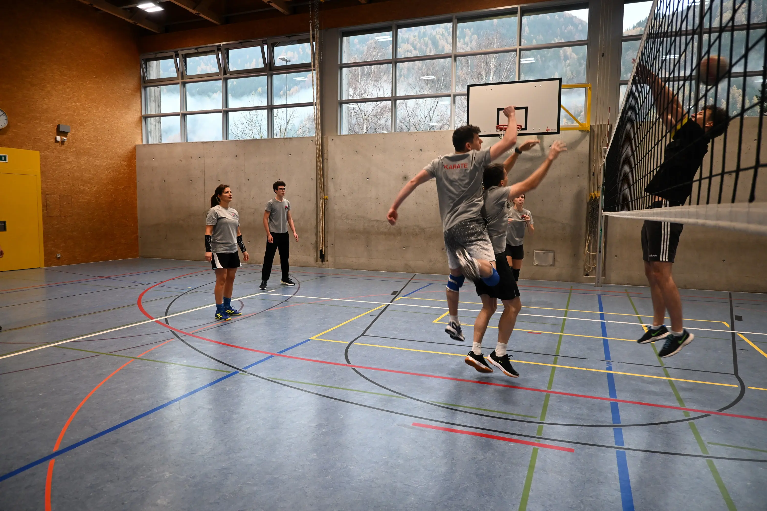 Un gruppo di ragazzi gioca a pallavolo in un campo coperto. Alcuni stanno saltando per colpire il pallone mentre altri osservano. Sullo sfondo si intravede un tabellone e finestre con vista verso l'esterno.