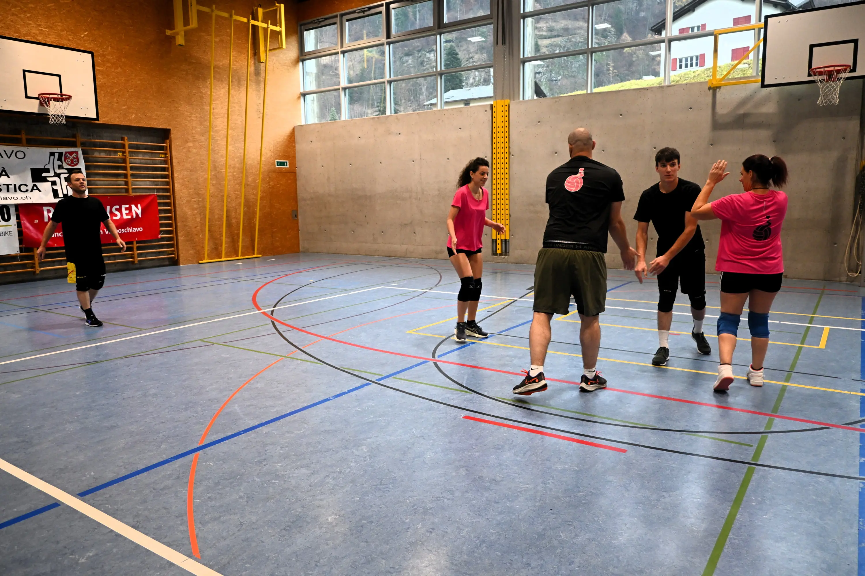 Un gruppo di persone gioca a pallavolo in un palazzetto. Due giocatori si preparano a ricevere la palla, mentre altri due sono in azione. Le pareti sono grigie e ci sono canestri di pallacanestro sullo sfondo. I