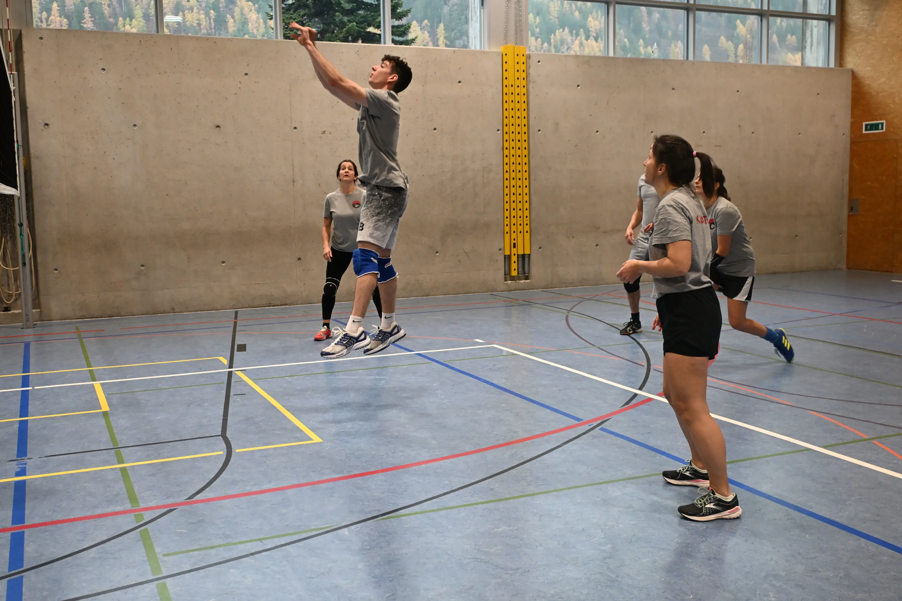 Giocatore di pallavolo mentre salta per una schiacciata, con altri tre compagni di squadra in campo. Il pavimento della palestra è segnato da linee colorate e si vedono finestre grandi sullo sfondo.
