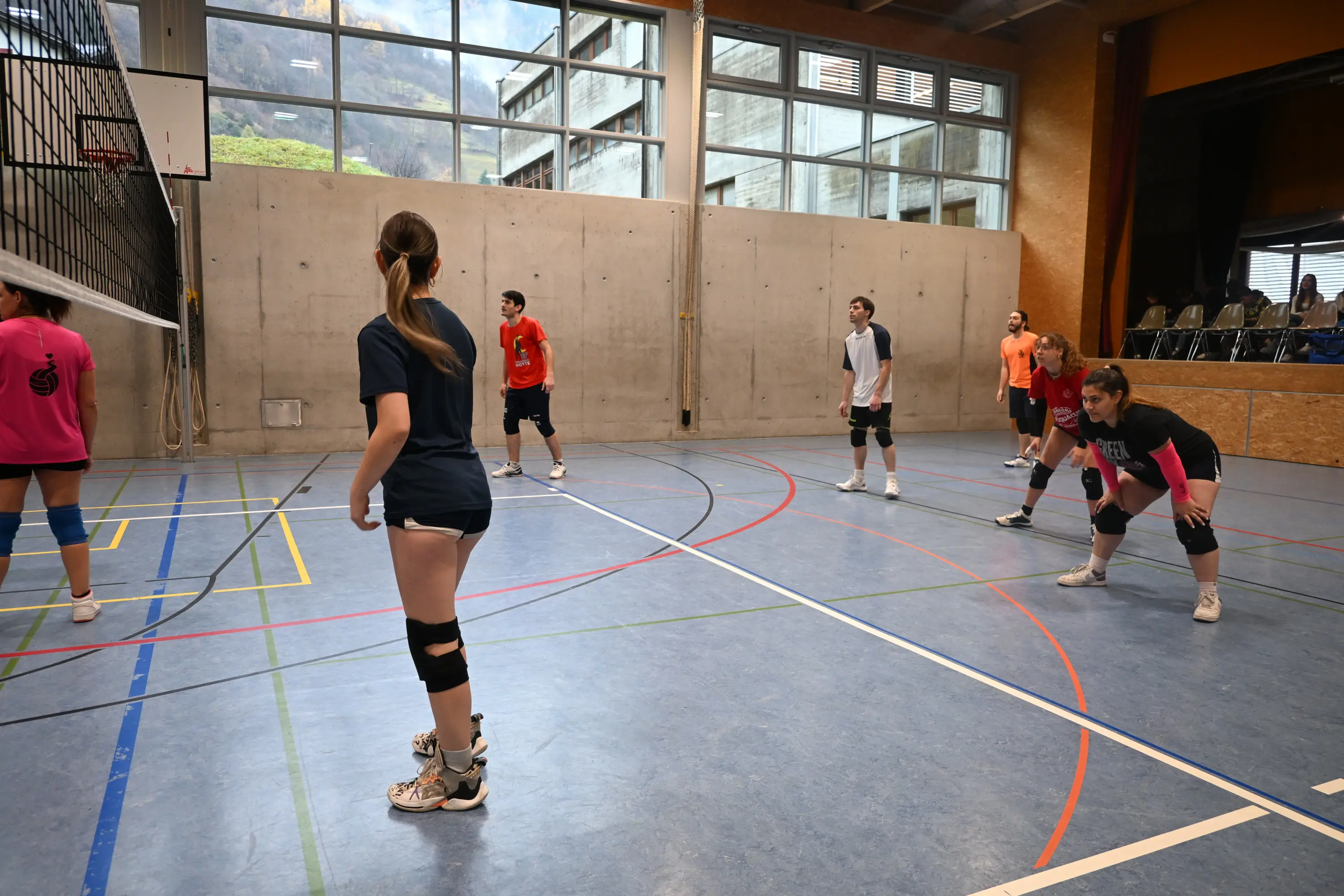 Giocatori di pallavolo in una palestra, pronti per una partita. Alcuni indossano magliette colorate e ginocchiere. Sullo sfondo, si vedono finestre grandi e sedie.