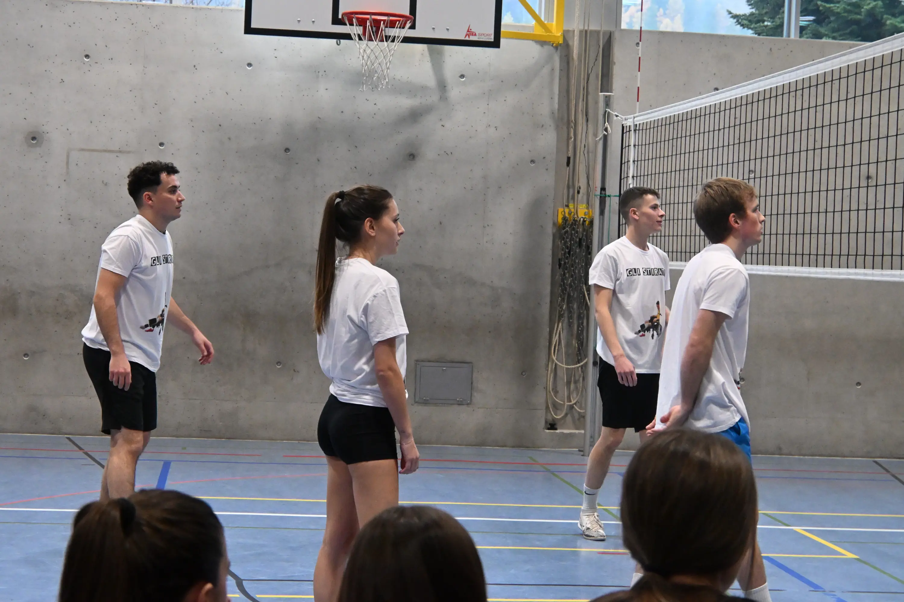 Quattro giocatori di pallavolo in attesa di ricevere il servizio, indossano magliette bianche e pantaloni sportivi. Sullo sfondo, una rete da pallavolo e un campo sportivo interno.