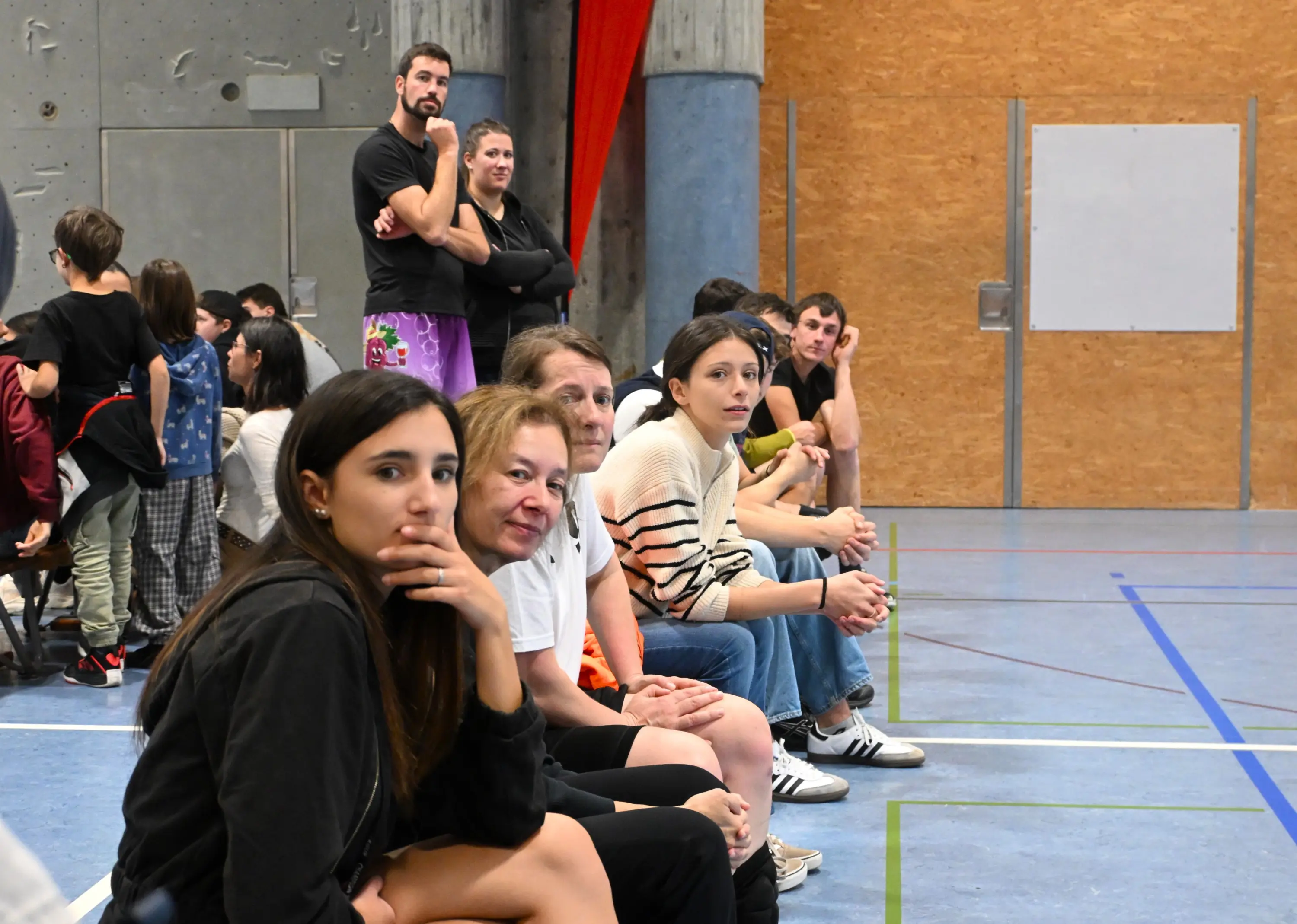Un gruppo di persone sedute in una palestra, alcune sorridono mentre altre osservano attentamente. Si notano giovani e adulti, con diversi stili di abbigliamento. Sullo sfondo, un muro attrezzato per arrampicata.