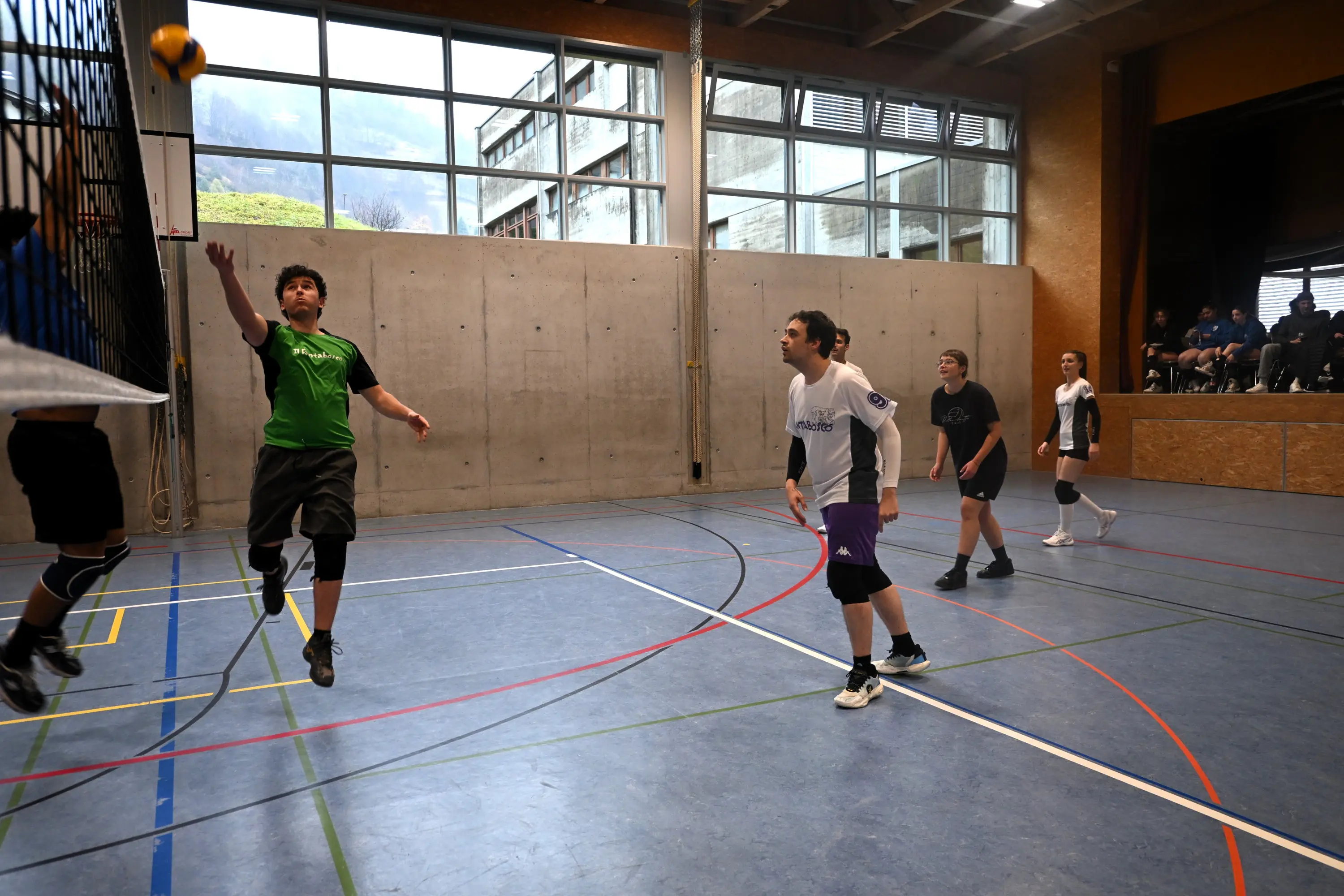 Giovani atleti giocano a pallavolo in un palazzetto. Un giocatore in maglia verde tenta di colpire la palla, mentre altri due sono pronti a riceverla. In fondo, si intravedono spettatori seduti.