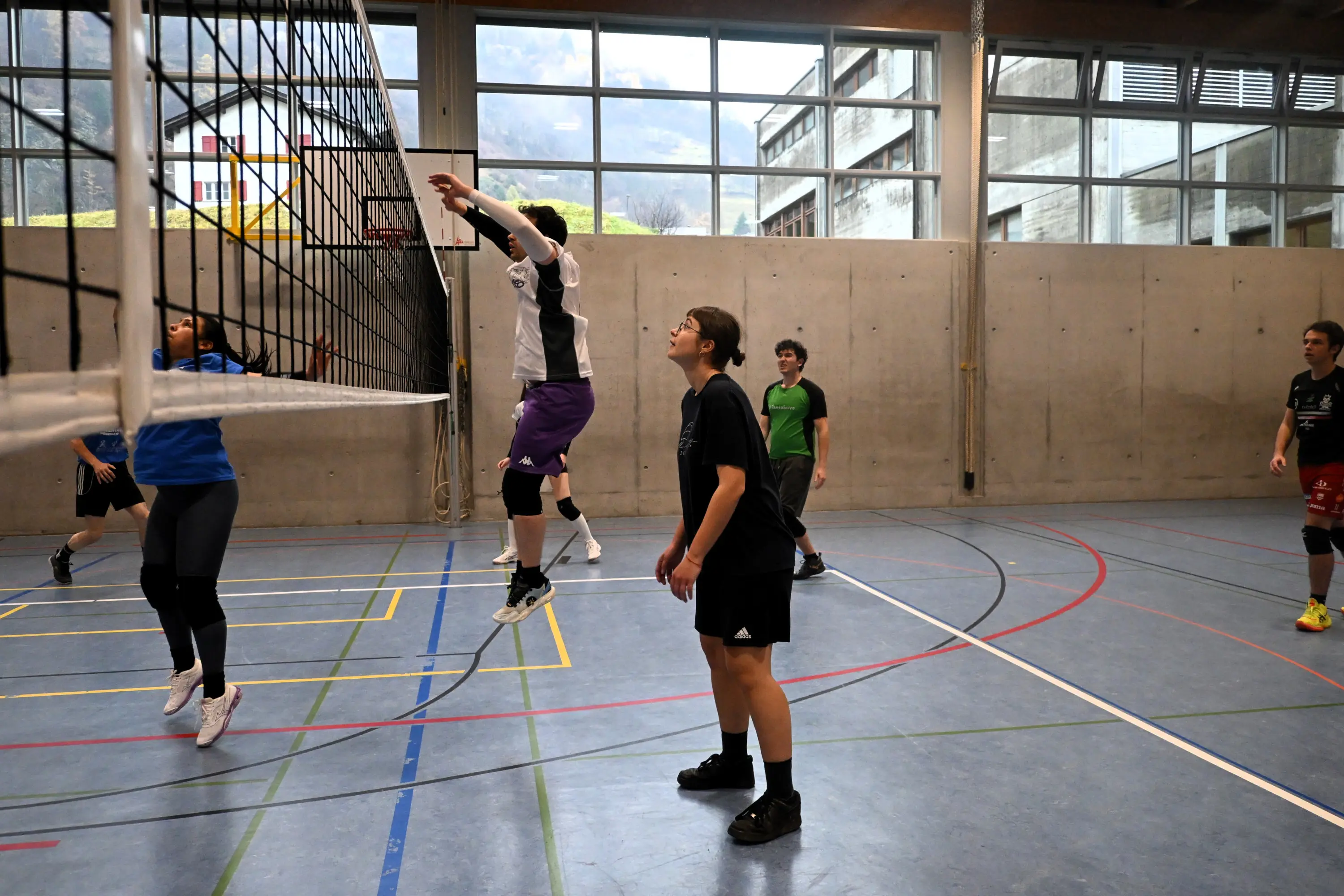 Giovani atleti che giocano a pallavolo in un campo al chiuso. Un giocatore salta per colpire la palla sopra la rete, mentre gli altri sono in attesa nelle loro posizioni.