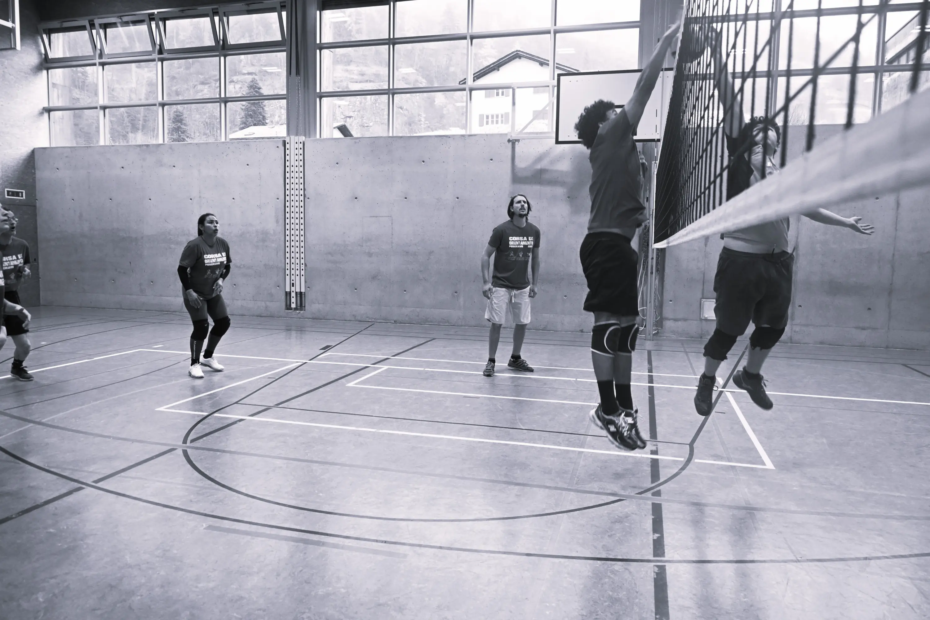 Giocatori di pallavolo in un campo al coperto, con due atleti che saltano per colpire la palla vicino alla rete. Alcuni membri del team indossano magliette coordinate. La luce naturale entra attraverso le finestre alte.