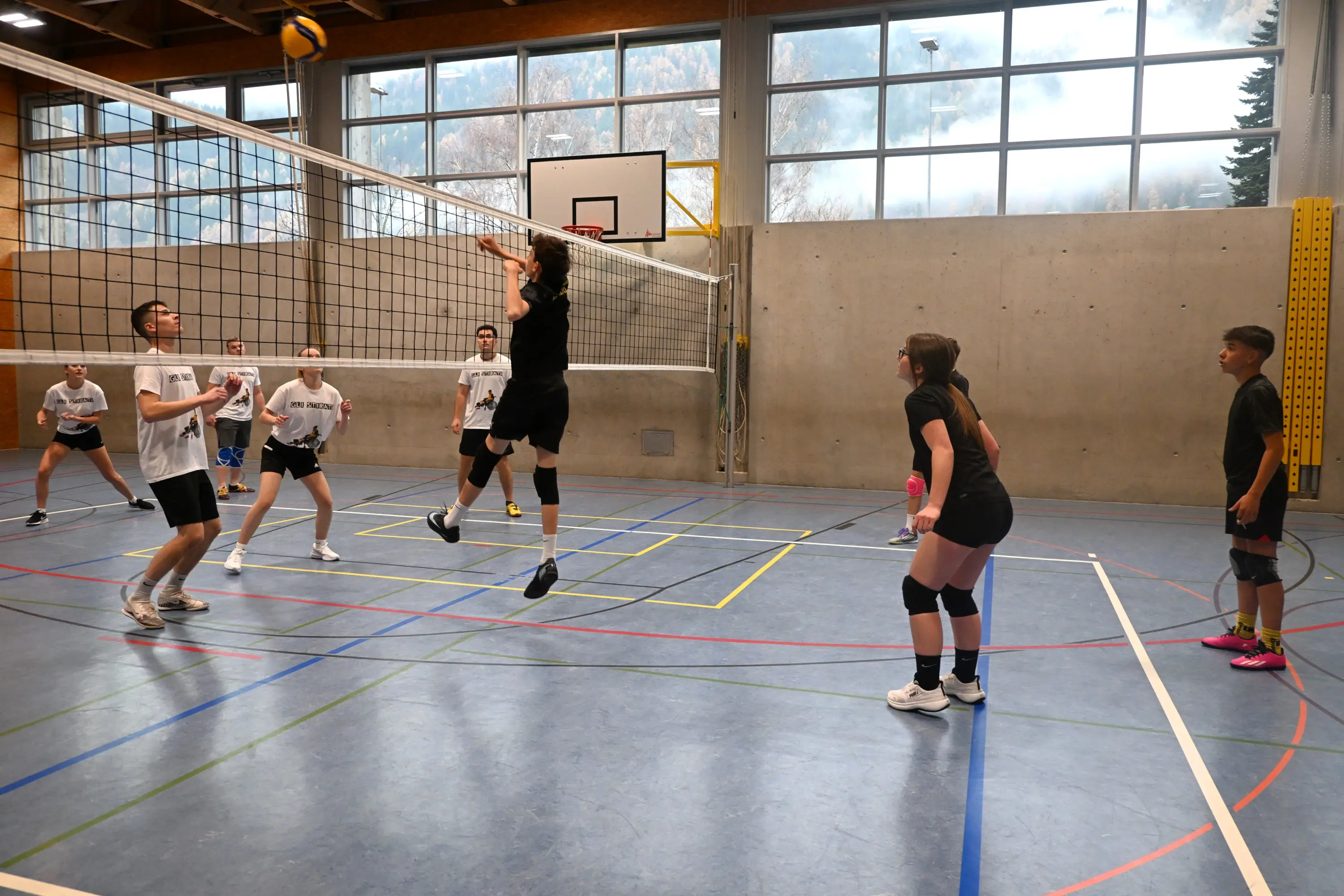Giocatori di pallavolo durante un allenamento in un campo indoor. Un atleta sta per colpire la palla sopra la rete mentre gli altri osservano e si preparano a reagire.
