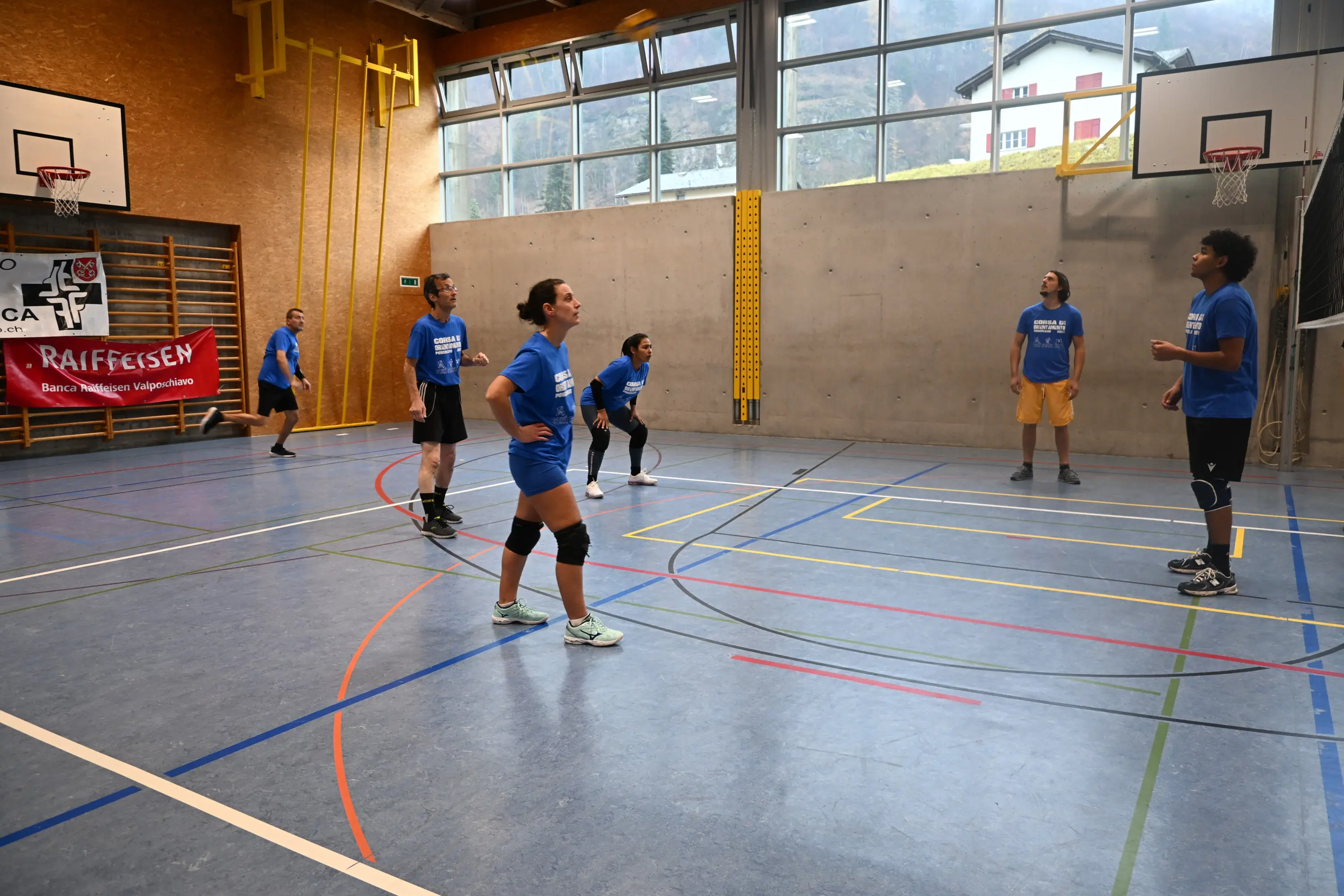 Un gruppo di sei persone si allena in un campo da pallavolo in una palestra, indossando magliette blu. Alcuni giocatori sono pronti a ricevere, mentre altri si preparano a colpire la palla. Sullo sfondo, si possono vedere le