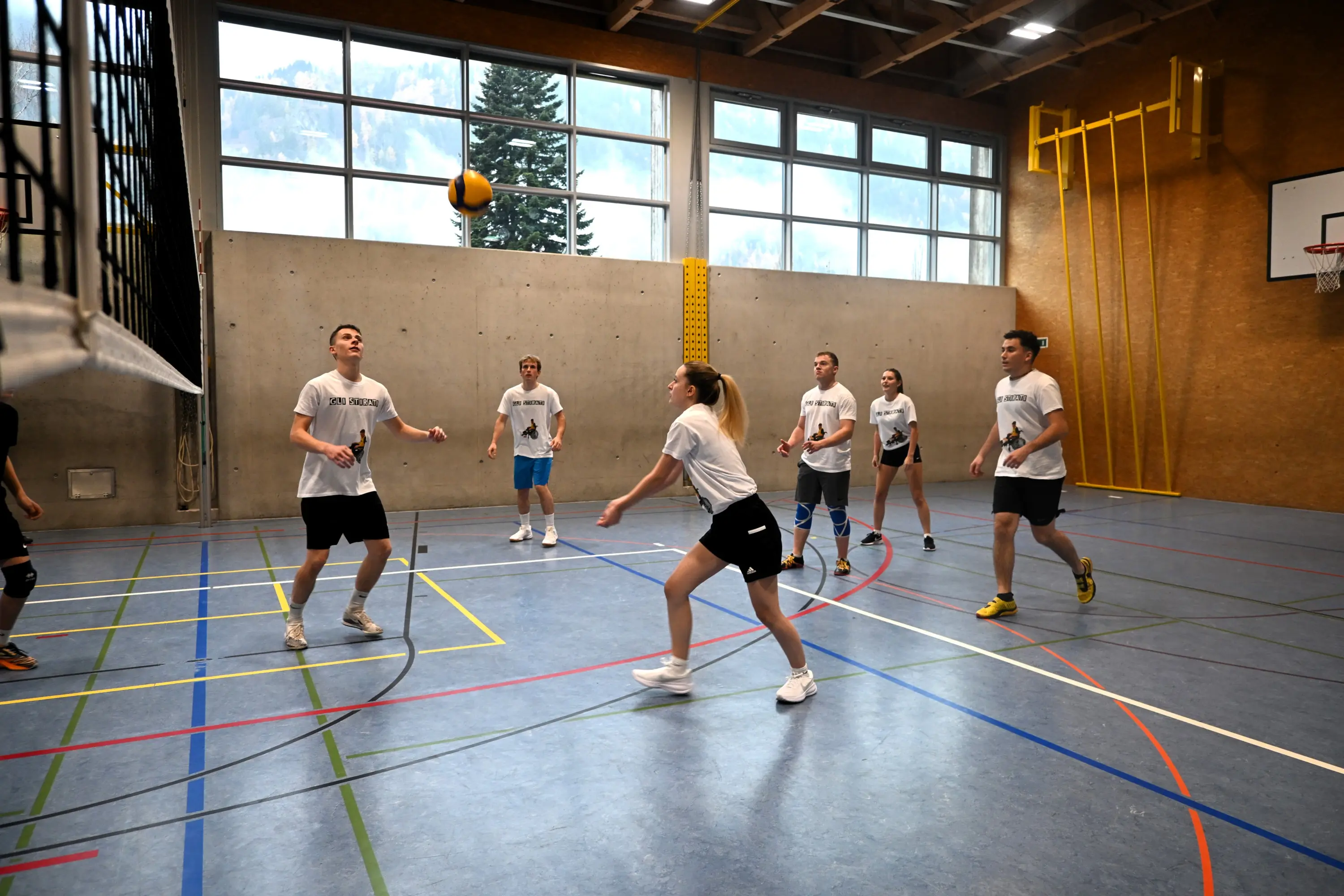 Un gruppo di giovani sta giocando a pallavolo in una palestra. Due ragazzi sono in attesa di ricevere la palla, mentre una ragazza sta per effettuare un passaggio. Le pareti sono chiare e ci sono finestre che mostrano alberi all'esterno.