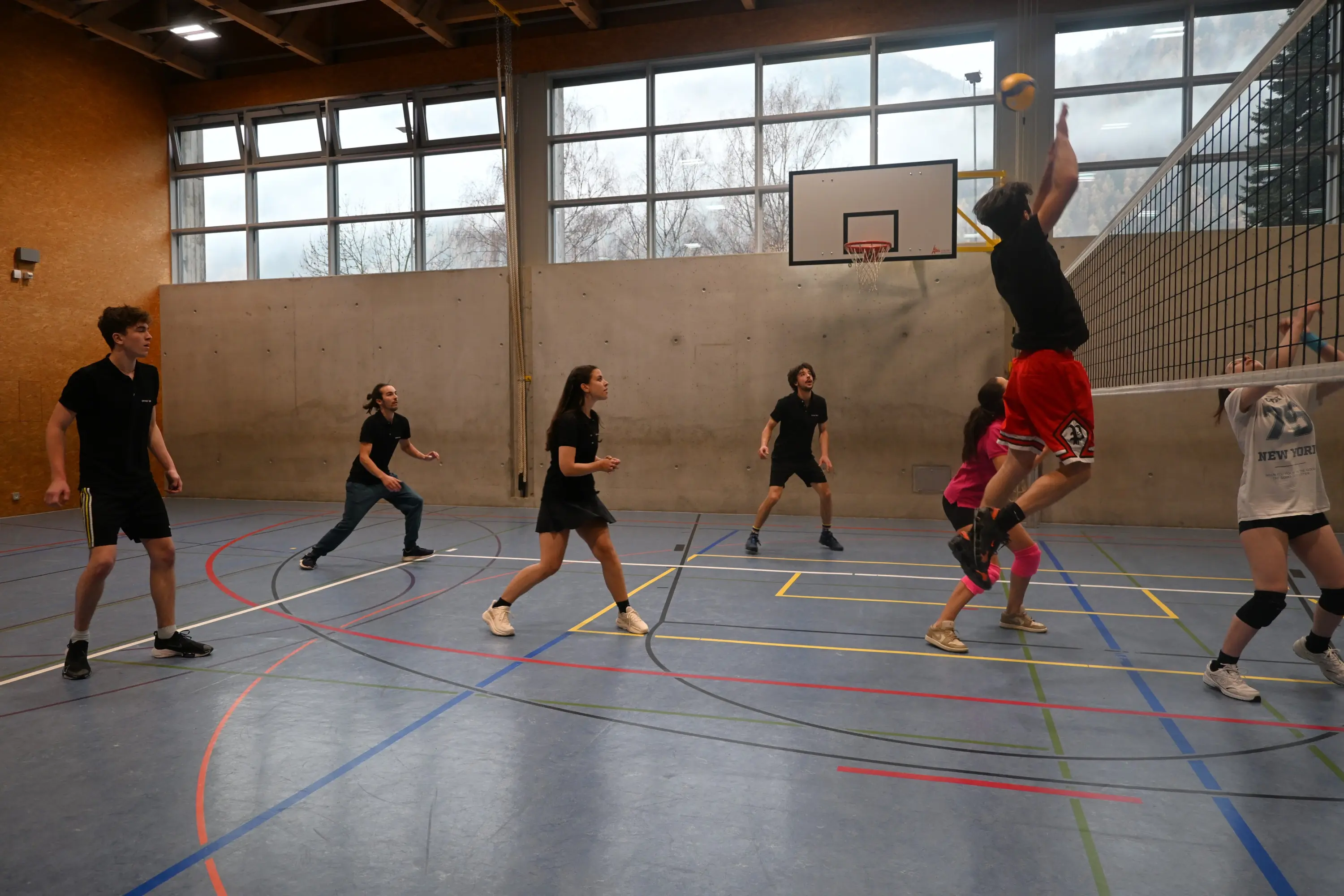 Un gruppo di giovani gioca a pallavolo in un campo al coperto. Un giocatore salta per colpire la palla sopra la rete, mentre gli altri si preparano per ricevere. Si notano diverse magliette e pantaloni sportivi.