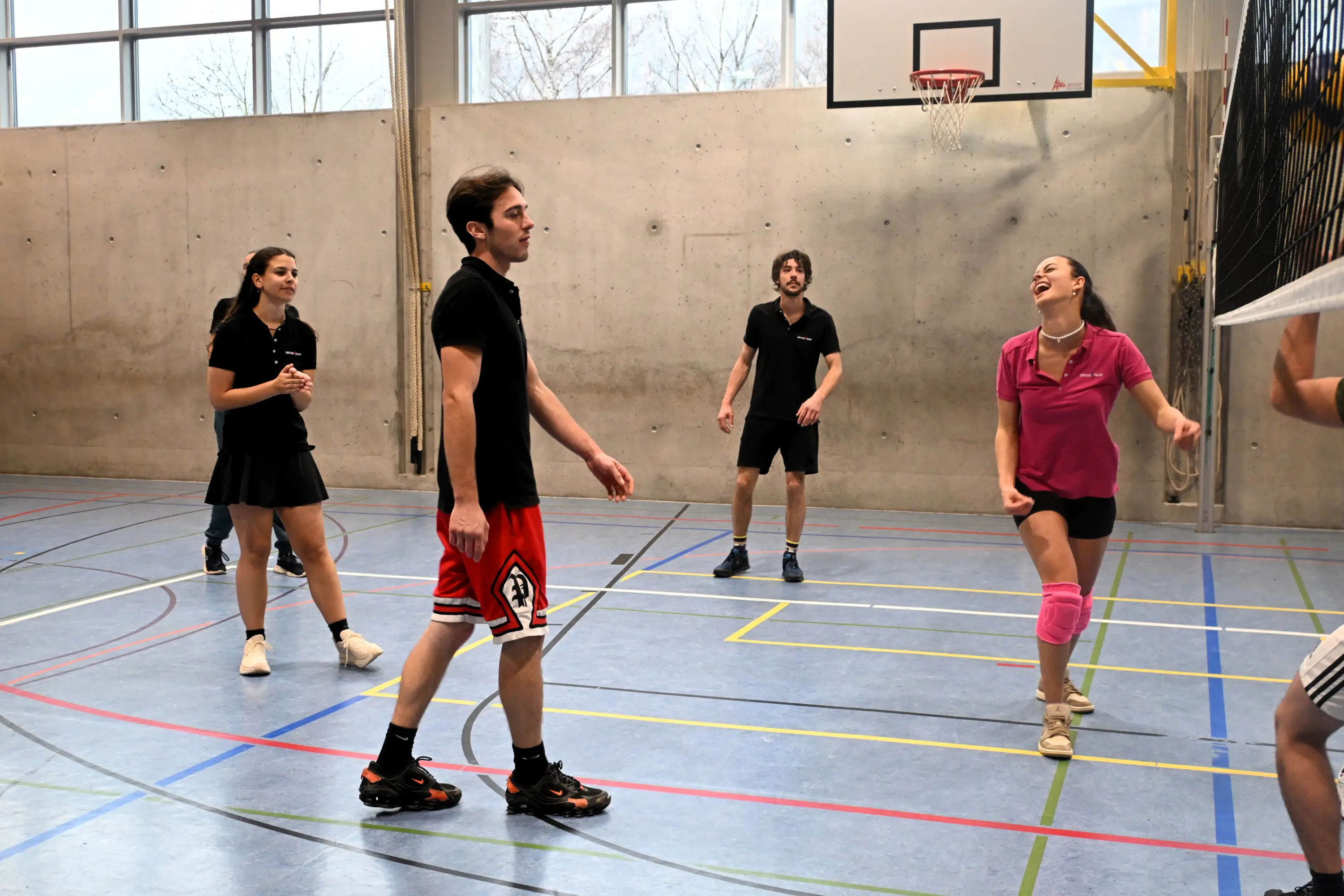 Un gruppo di giovani gioca a pallavolo in una palestra. Due ragazze indossano abbigliamento sportivo nero e rosa, mentre i ragazzi sono in t-shirt e pantaloni corti. Si vede una rete da pallavolo e un canestro sullo sfondo.