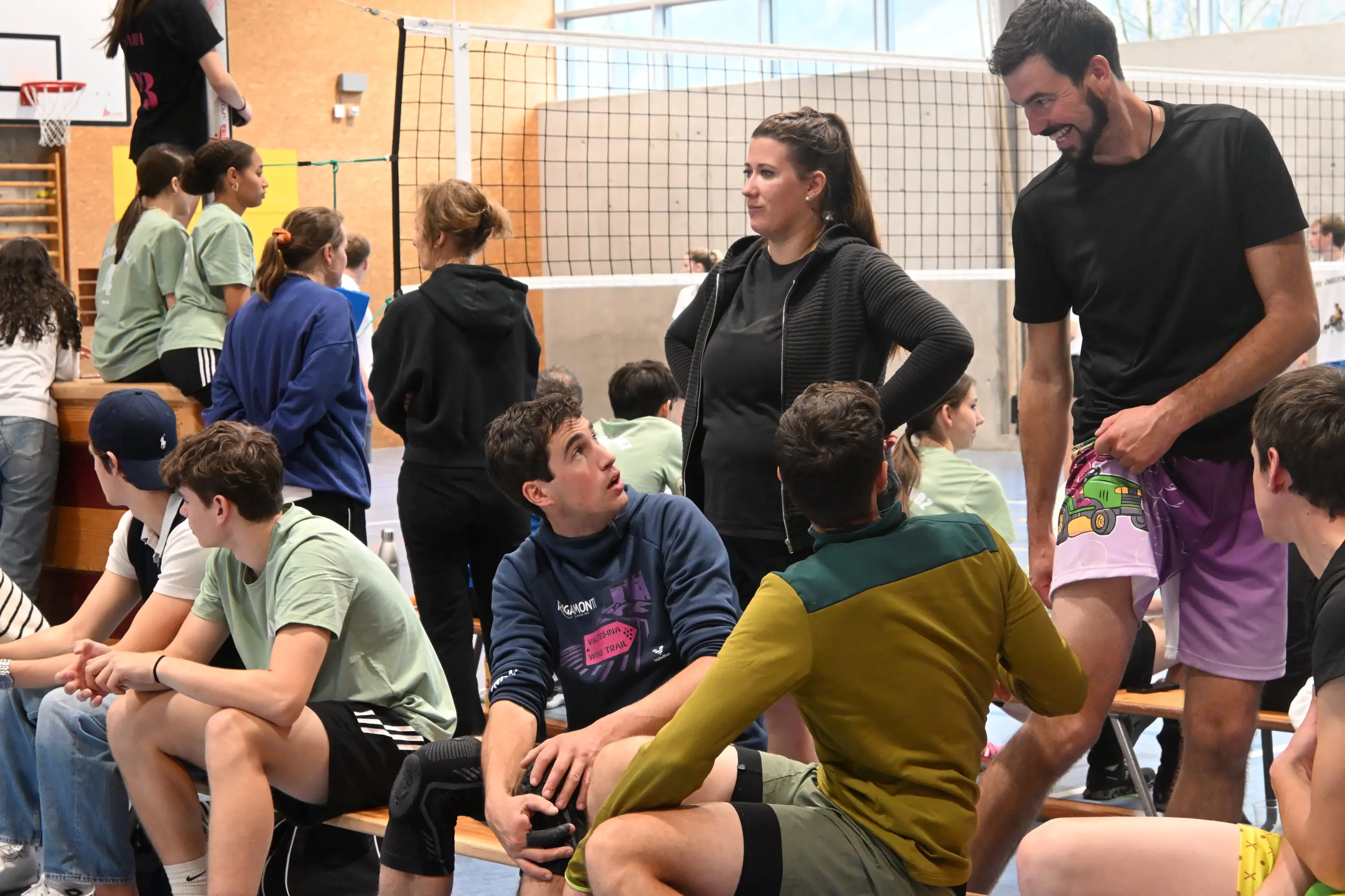 Un gruppo di giovani e adulti si riunisce in una palestra, alcuni seduti su panchine e altri in piedi. C'è una donna che conversa con un uomo mentre altri ragazzi li ascoltano con interesse. Sullo sfondo si vede una rete da pallavolo e diversi