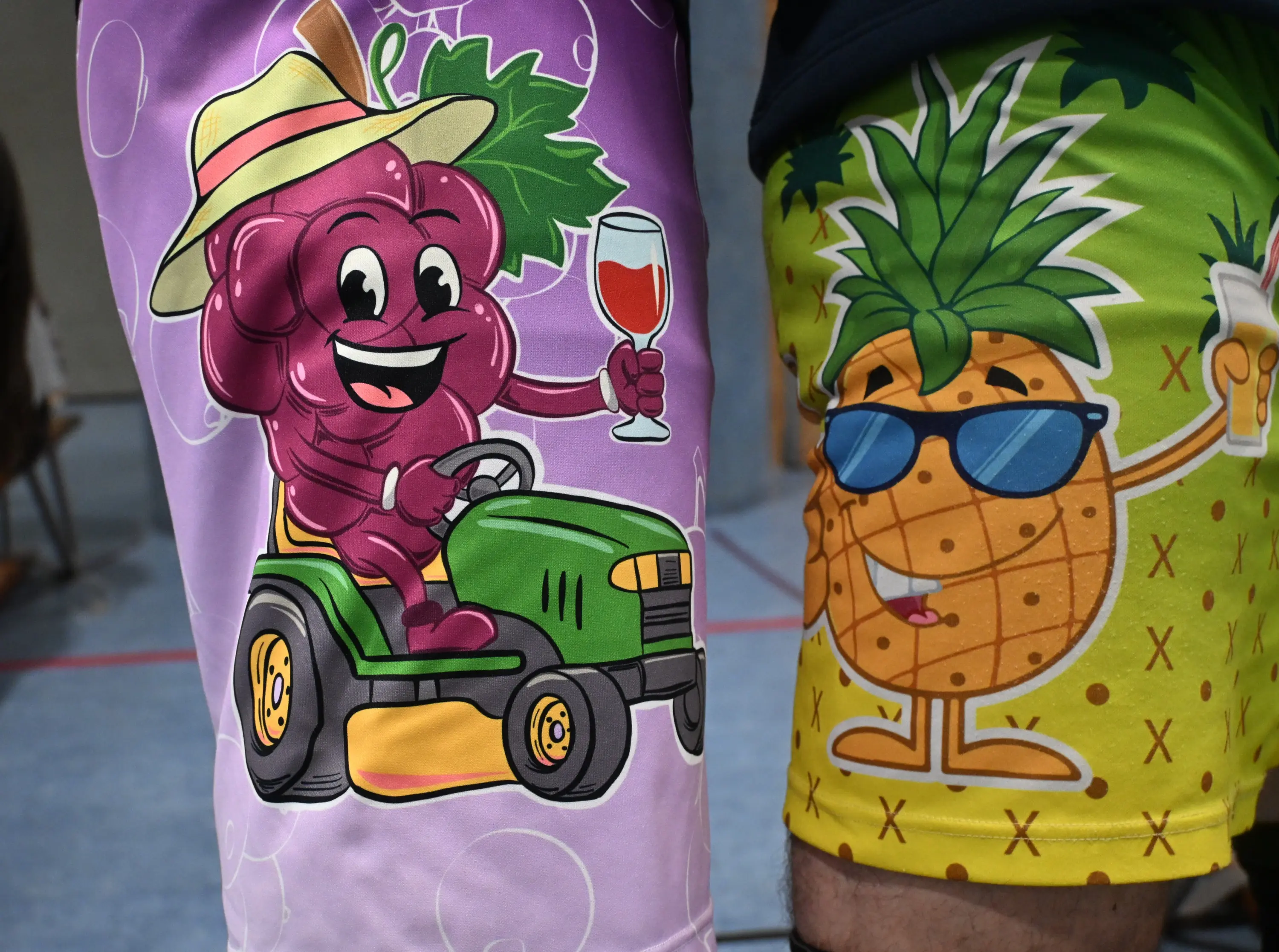 Pantaloni da bagno colorati, uno con un personaggio d'uva sorridente che guida un trattore e tiene un bicchiere di vino, l'altro con un'ananas occhialuta che sorseggia una bevanda.