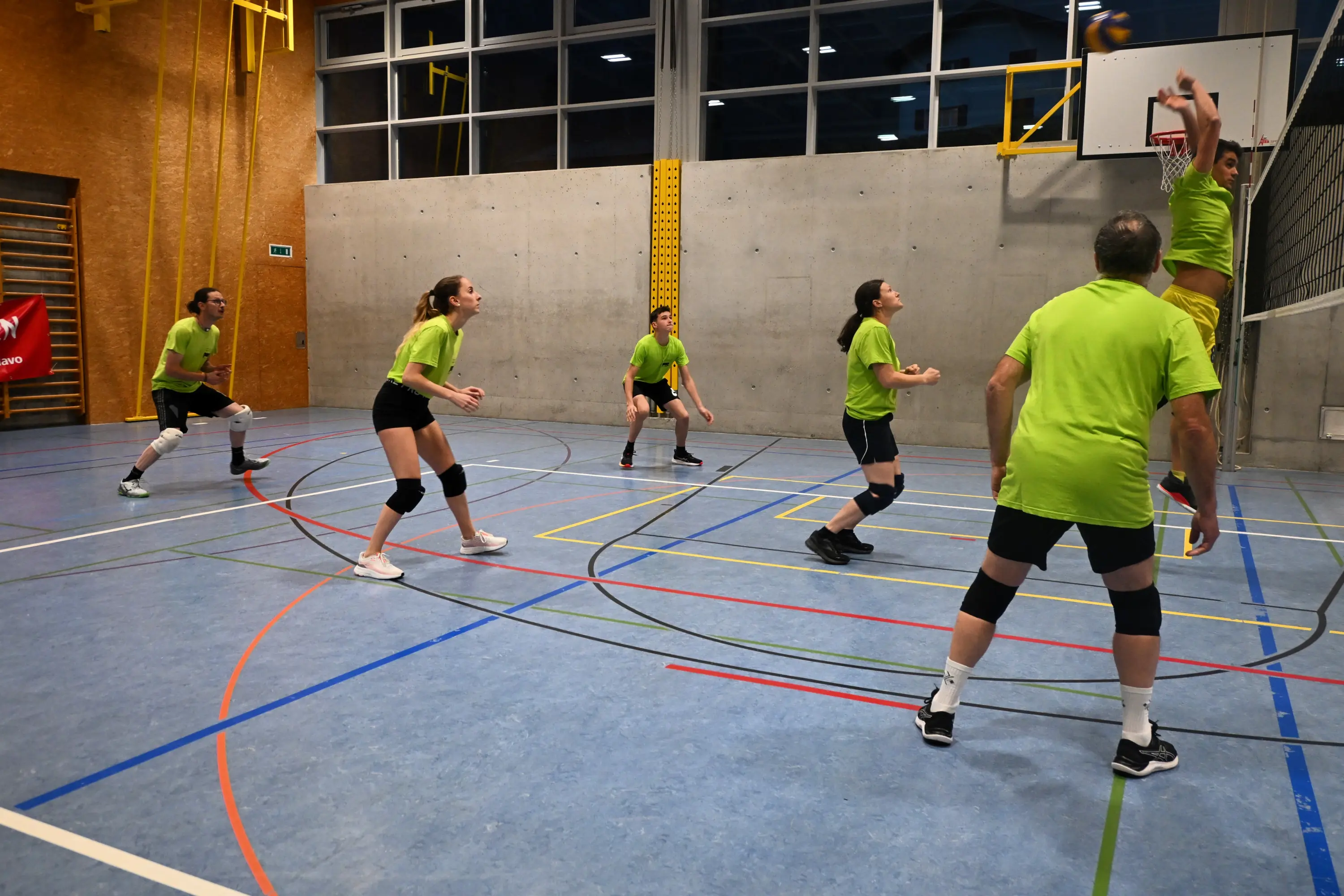Un gruppo di sei persone gioca a pallavolo in una palestra. Sono vestiti con magliette verdi e pantaloni corti neri. Un giocatore sta saltando per schiacciare la palla verso il canestro, mentre gli altri osservano attentamente.