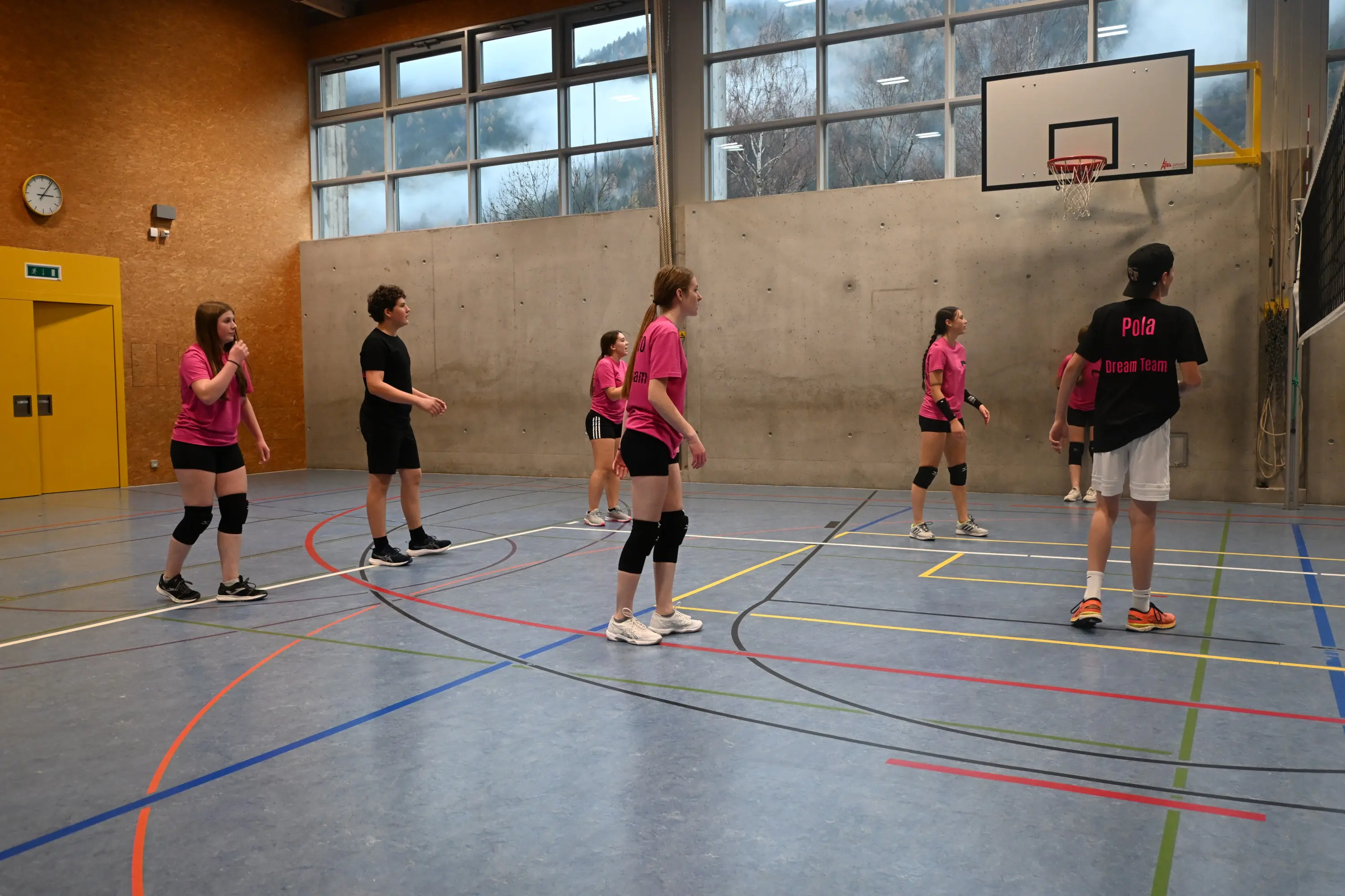 Un gruppo di giovani atlete gioca a pallavolo in una palestra. Indossano magliette rosa e pantaloni neri, mentre un giocatore è in prossimità della rete. Sullo sfondo ci sono finestre che mostrano un paesaggio montano.