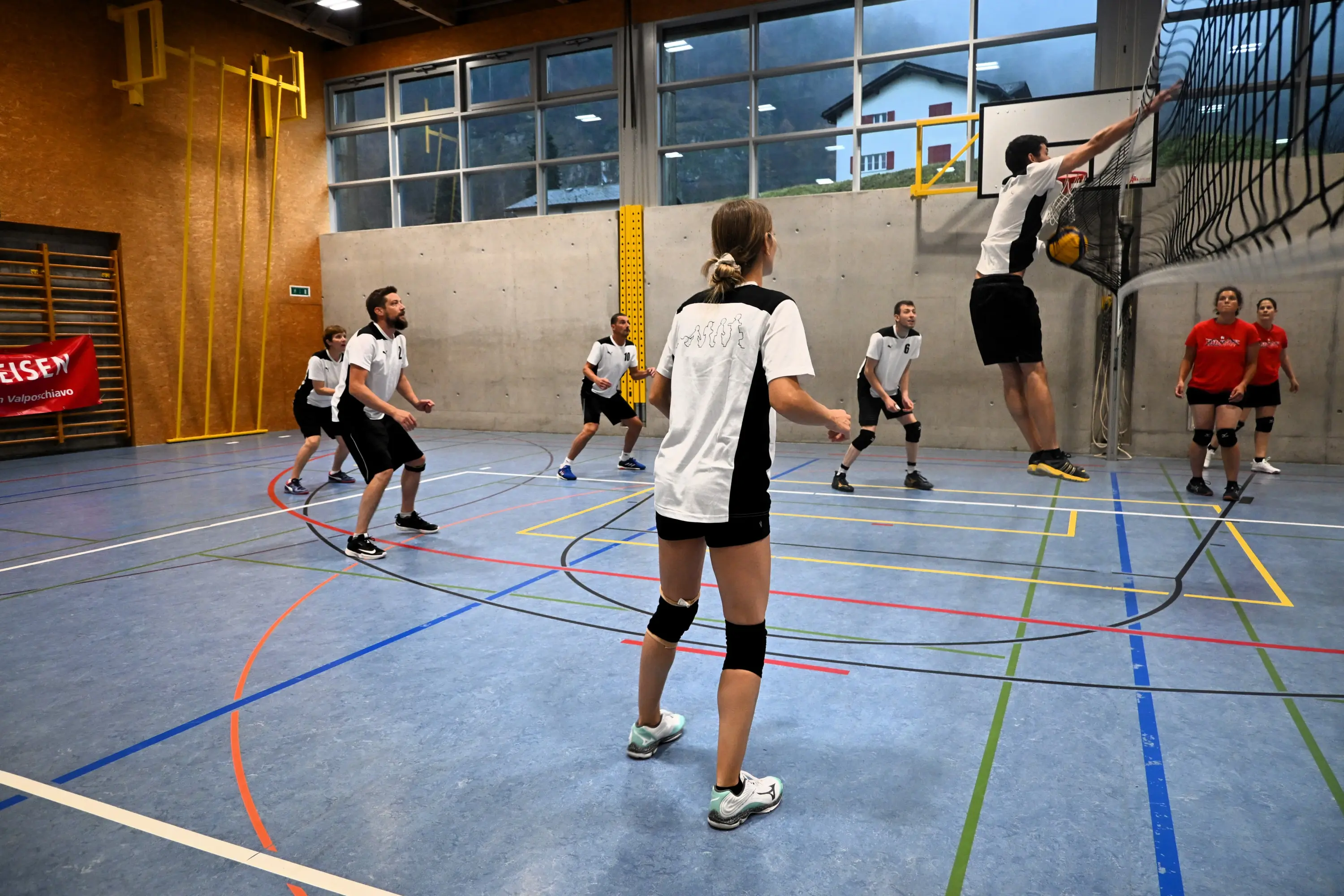 Giocatori di pallavolo in un campo al coperto, impegnati in una partita. Alcuni partecipanti sono in movimento, mentre uno salta per colpire la palla. I giocatori indossano magliette bianche e nere, con un'atmos
