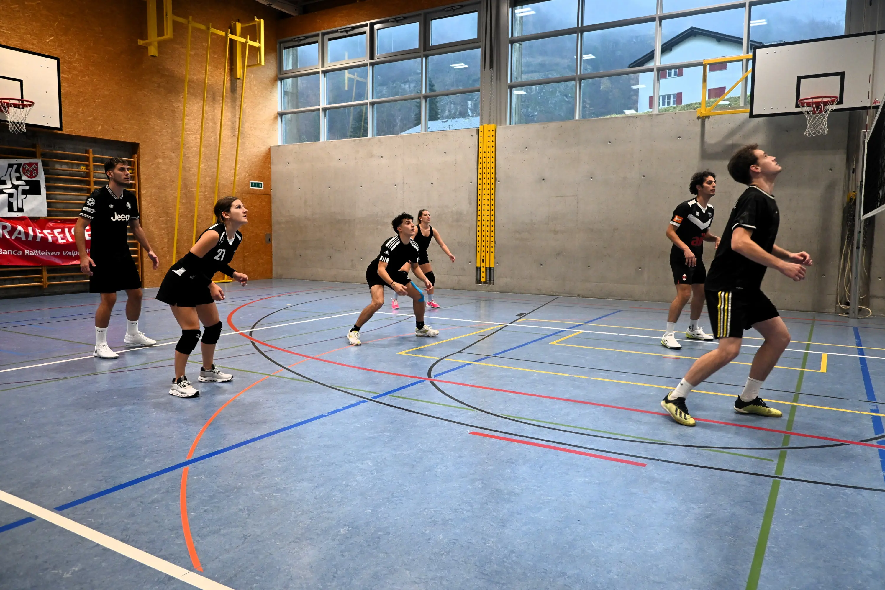 Un gruppo di sei giovani giocatori di pallavolo si allena in una palestra. Tre giocatori sono in posizione di attesa, mentre altri tre stanno per ricevere la palla. I ragazzi indossano magliette nere con dettagli bianchi e sono concentrati sulla sessione di