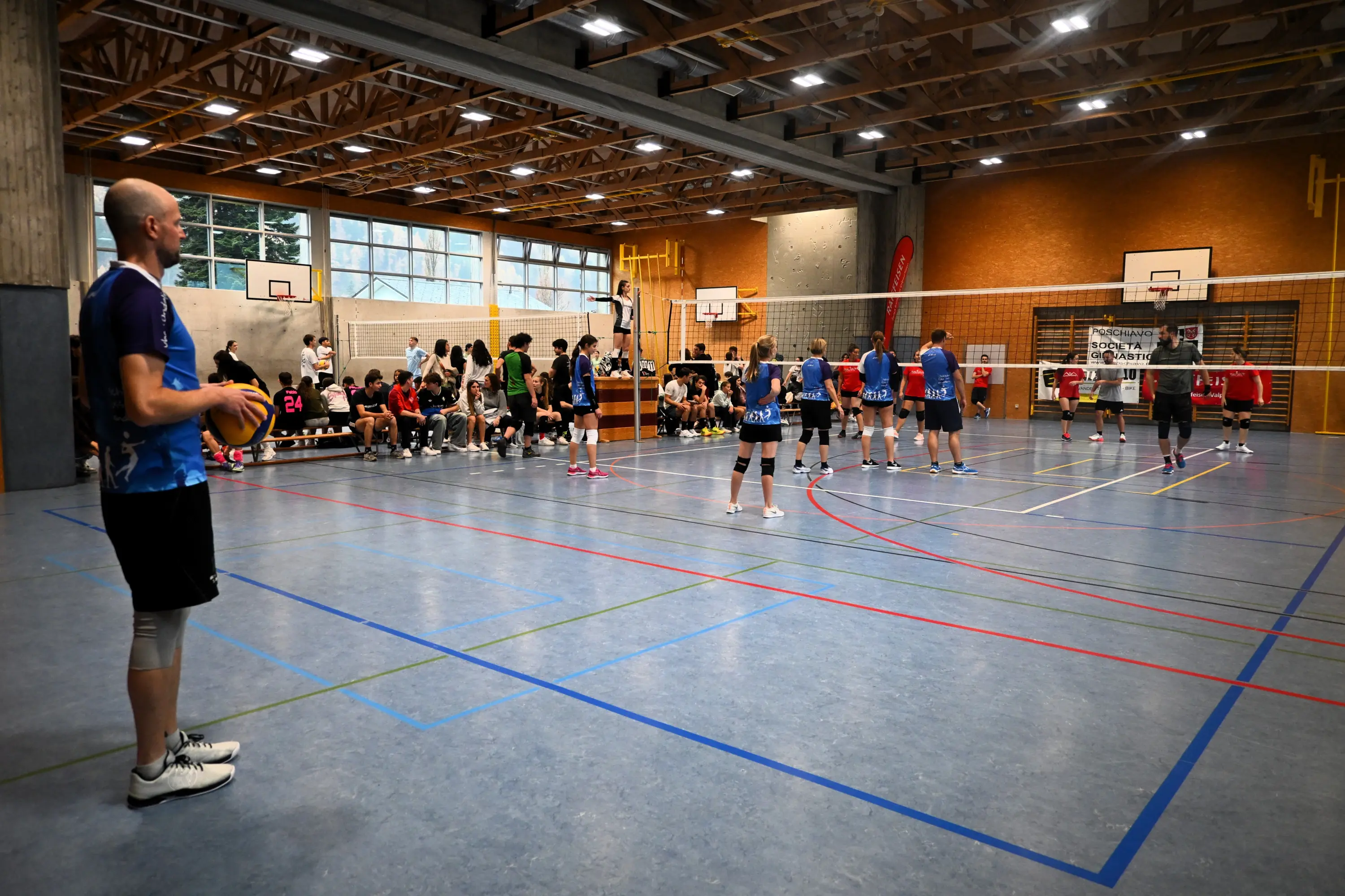Un campo da pallavolo all'interno di un centro sportivo. Giocatori in azione, mentre un allenatore osserva dalla sideline. Il pubblico è seduto lungo le tribune laterali.