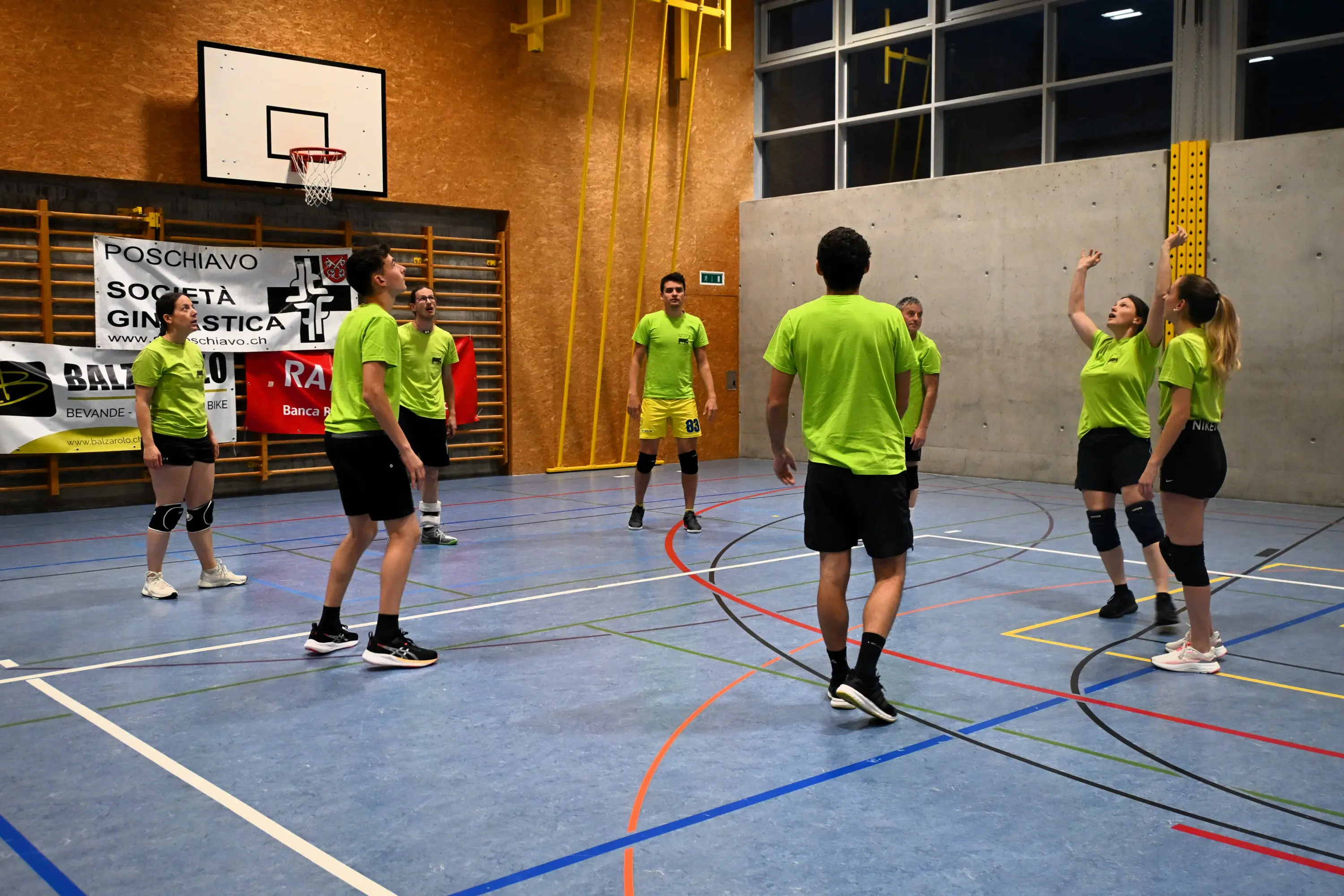 Un gruppo di giovani in magliette verdi vivaci si allena in una palestra. Alcuni giocatori si preparano a colpire la palla mentre altri osservano. Sullo sfondo, un banner con il logo di una società sportiva.