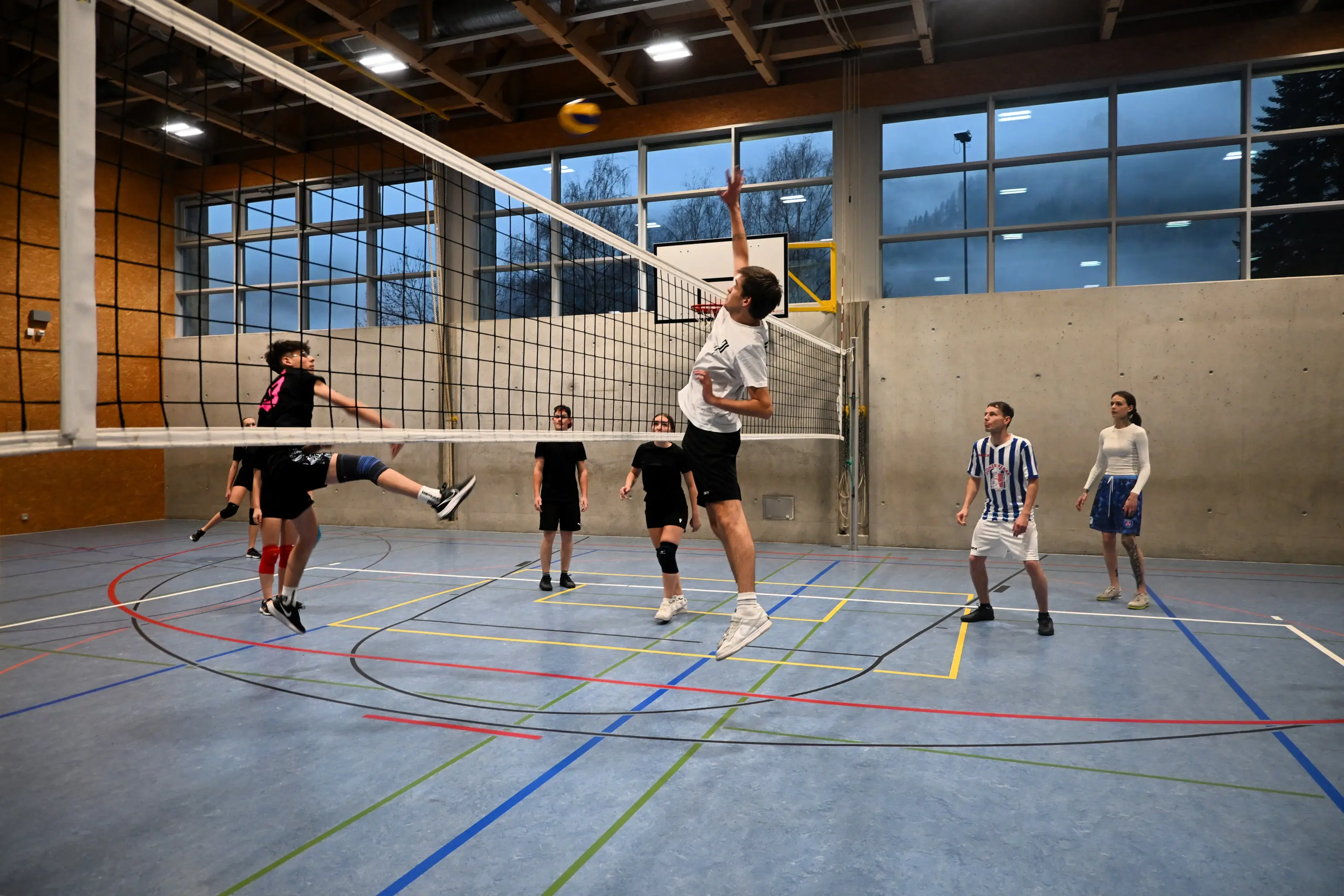 Giocatori di pallavolo nel momento di un attacco, con uno in salto che colpisce la palla sopra la rete. Altri giocatori si trovano nelle diverse posizioni del campo, in una palestra con finestre che permettono l'ingresso della luce.