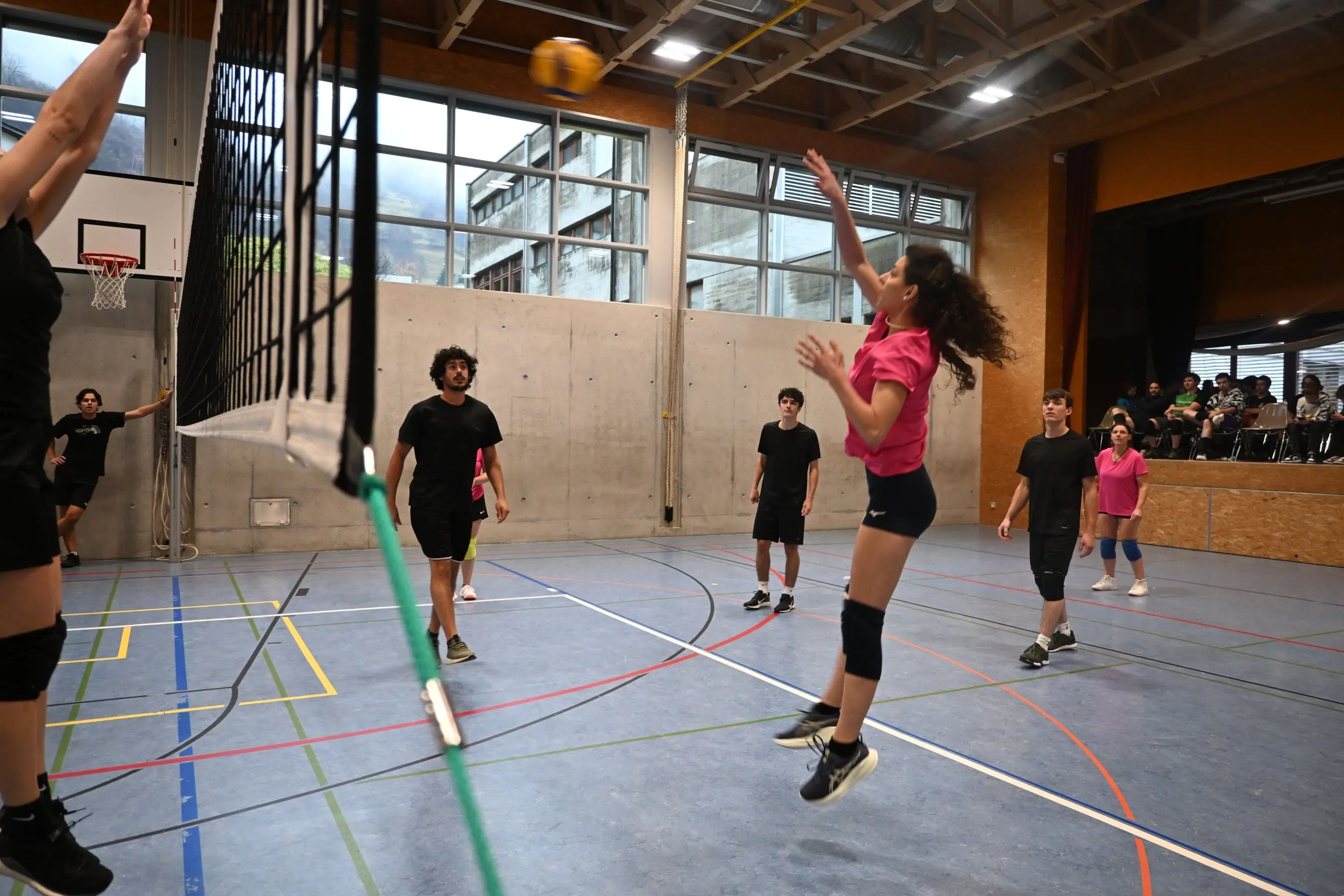 Giovani giocatori di pallavolo in una palestra. Una ragazza con una maglietta rosa salta per colpire il pallone sopra la rete, mentre altri giocatori osservano.
